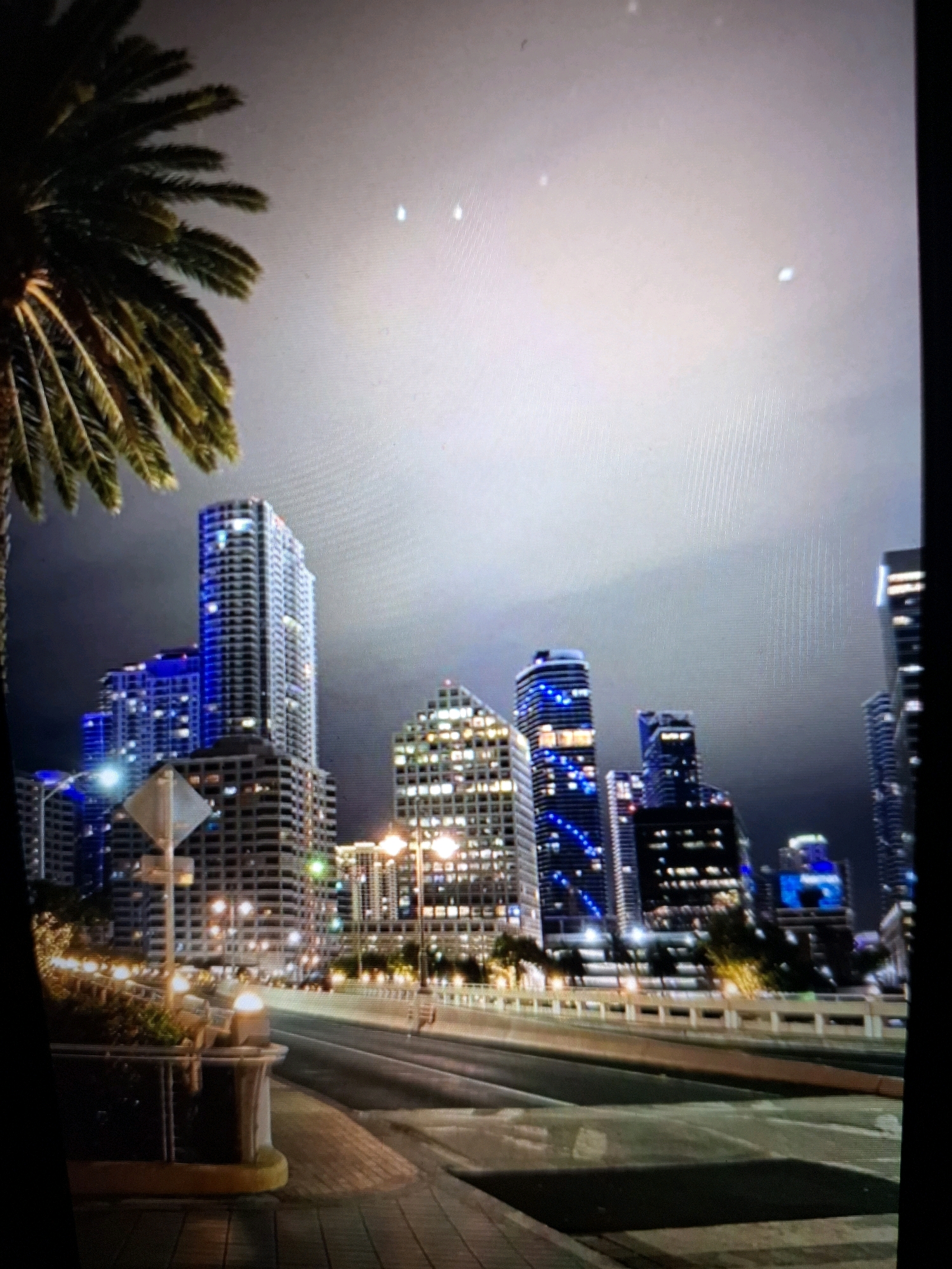The image displays a nighttime cityscape with several tall buildings illuminated by lights. A palm tree is partially visible in the foreground on the left side. The buildings feature various architectural styles and window patterns, with some exhibiting blue lighting. A road is visible in the lower part of the image, with streetlights and what appears to be a pedestrian walkway. The sky is overcast and dark. There are no people visible in the image. The scene is likely an urban environment. The location of Yamunanagar, India is not depicted in this image.