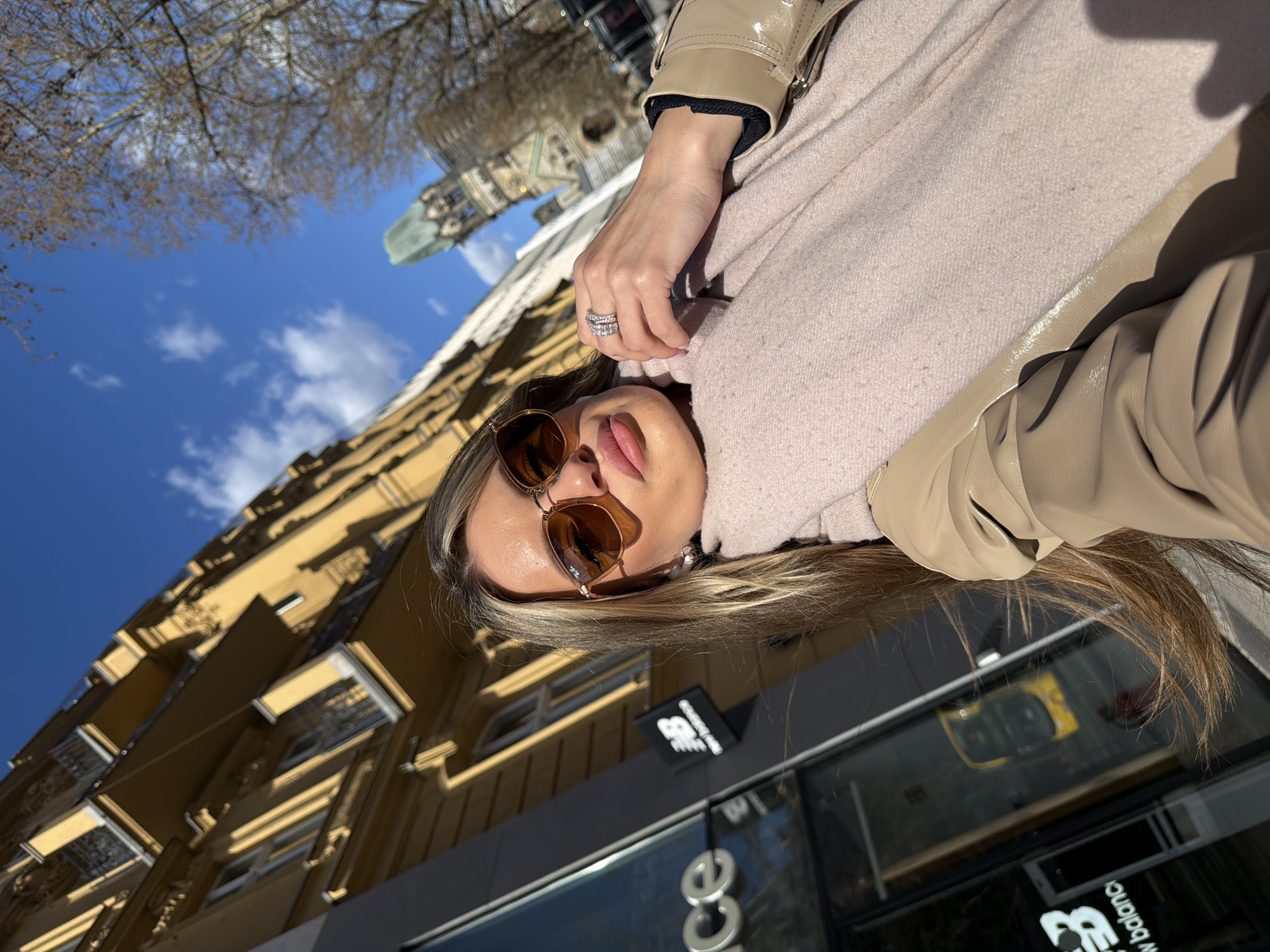A low-angle, tilted photograph shows a woman wearing brown sunglasses, a light pink scarf, and a beige jacket. She is wearing a ring on her hand. Behind her is a yellow building with windows and balconies. Part of a taller building with a green dome can be seen. A business with the words "new balance" is partially visible. The photograph appears to have been taken in Berlin, Germany, near the Neue Kirche church.