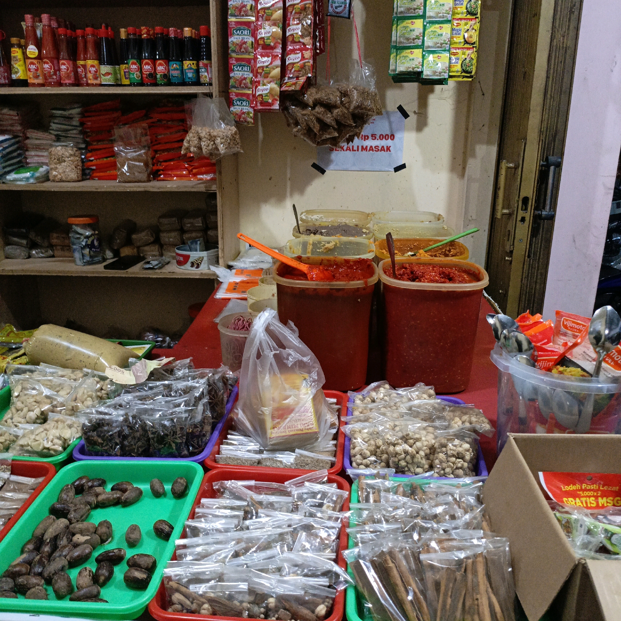 The image depicts a street food stall in Salatiga, Indonesia, specializing in spices and condiments. The stall is organized with various items displayed on shelves and in trays on a red table. On the shelves, there are rows of bottled sauces and sachets of spices. The table is laden with an assortment of goods, including large containers of red chili paste, bags of nuts, star anise, cardamom, cinnamon sticks, and other dried spices. Some items are packaged in clear plastic bags, while others are loose in trays or jars. A prominent sign reads "Rp 5.000 Sekali Masak", indicating the price and purpose of some of the products. A cardboard box is also visible, containing sachets of MSG. The lighting suggests an indoor setting, possibly a market or a covered stall, with natural light coming from the right side of the image. No people are present in the frame.