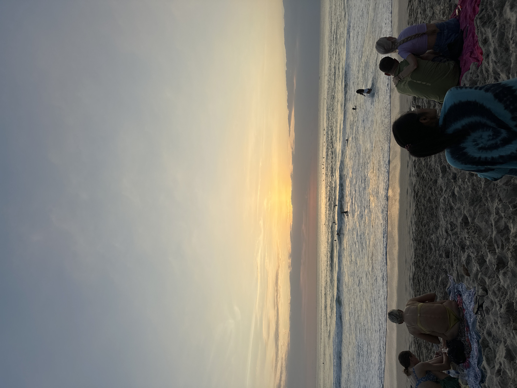 The image depicts a serene beach scene in Kuta Selatan, Indonesia, during sunset. The sky is painted with soft hues of orange and yellow, transitioning to a pale blue at the top. Wisps of clouds add texture to the sky. The ocean is calm with gentle waves rolling onto the sandy shore. Several surfers are scattered across the water, enjoying the evening surf.
In the foreground, a group of people are seated on the sand, facing the ocean. On the left, two individuals are sitting together, seemingly engaged in conversation or relaxing. To their right, a couple is embracing, with one person's arm around the other's shoulder. Further to the right, another person is seated facing away from the camera, also observing the sunset. The sand is covered in footprints, indicating it's a popular spot. Beach towels and personal belongings are visible on the sand, adding to the casual atmosphere. The overall impression is one of tranquility and enjoyment of nature's beauty.