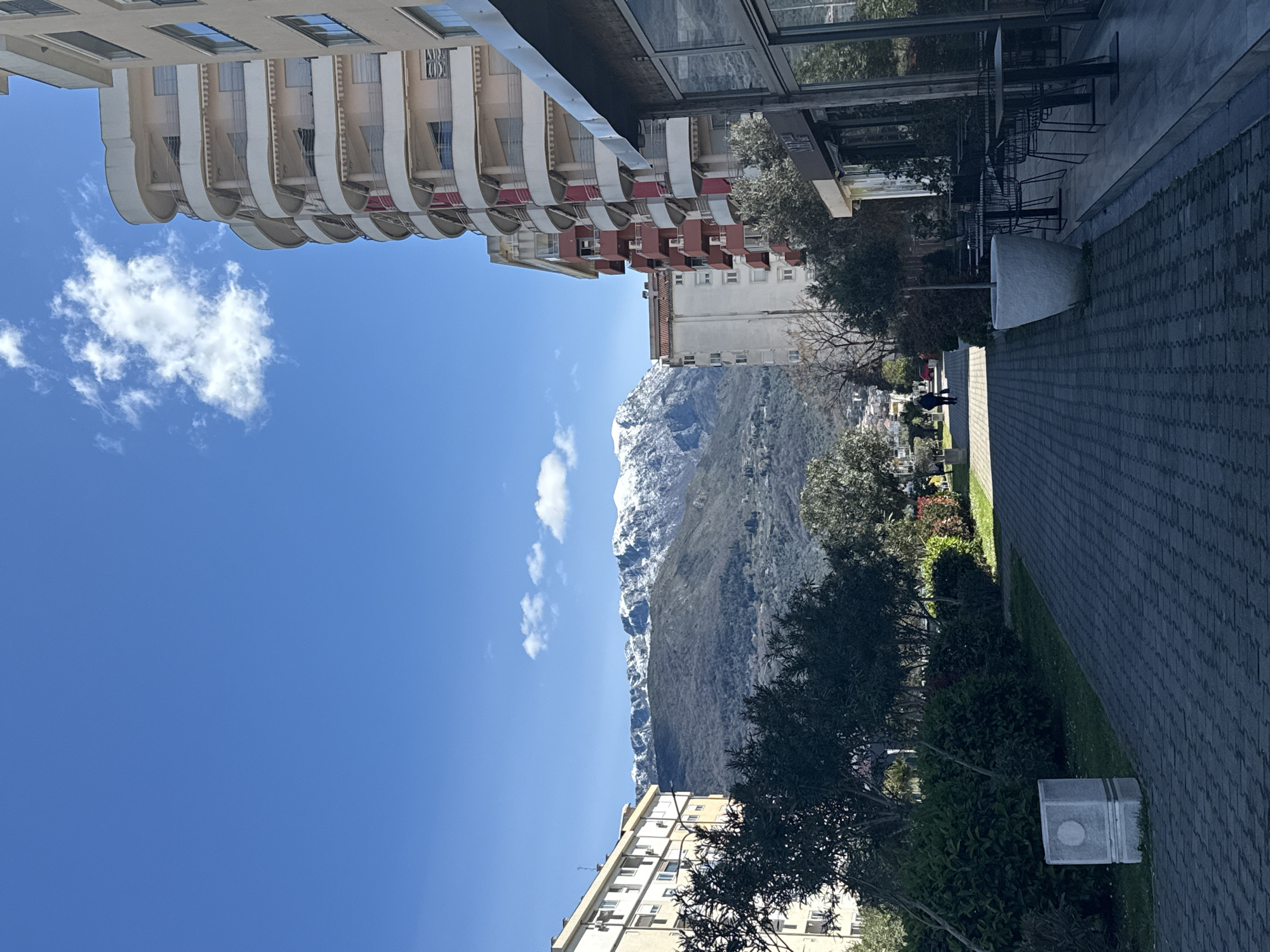 The image shows a street scene in Bar, Montenegro, with a view of the Rumija mountain range in the background. The sky is clear and blue, with a few scattered clouds. On the left side of the image, there are apartment buildings, one with white balconies and red accents, and another one further down. On the right side, there is a paved sidewalk lined with trees and bushes. The outdoor seating area of a restaurant or cafe is also visible, with tables and chairs. The Rumija mountains are visible in the distance, with snow-capped peaks.