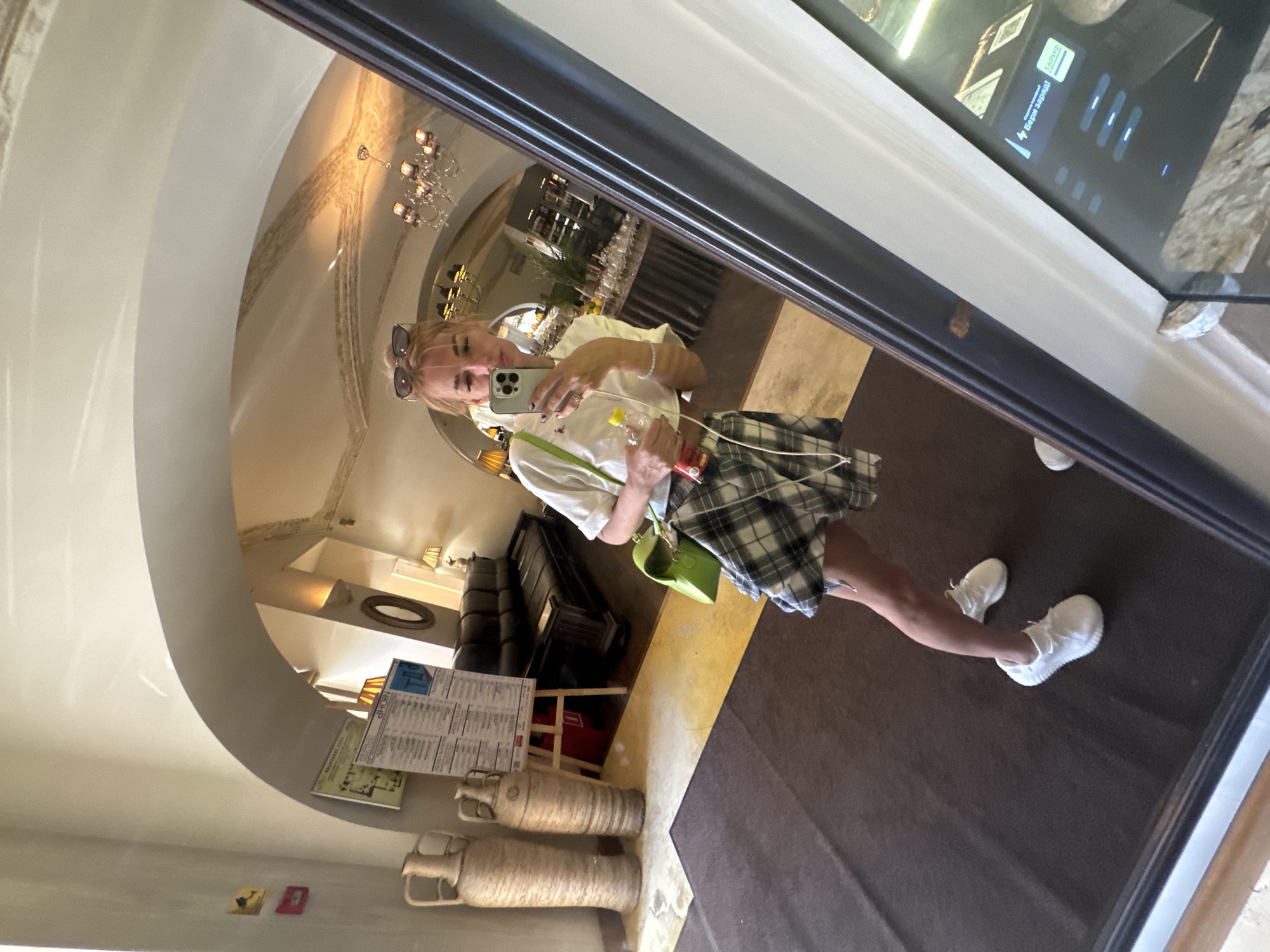 A young woman with blonde hair pulled back and sunglasses perched on her head takes a mirror selfie in what appears to be a hotel lobby or a restaurant. She is wearing a white cropped t-shirt, a grey and black plaid skirt, and white sneakers. She holds a smartphone in her right hand and a bottle with a red label and yellow cap in her left hand. A bright green cross-body bag hangs from her left shoulder. In the background, reflected in the mirror, are large decorative vases, a black leather sofa, a chandelier, and a bar area with bottles. The architecture suggests an indoor space with arched ceilings. The digital display on the right side of the image shows text in Russian, including "БЕРИ ЗАРЯД!" and "ТАРИФ", indicating the location is likely in Saint Petersburg, Russia.