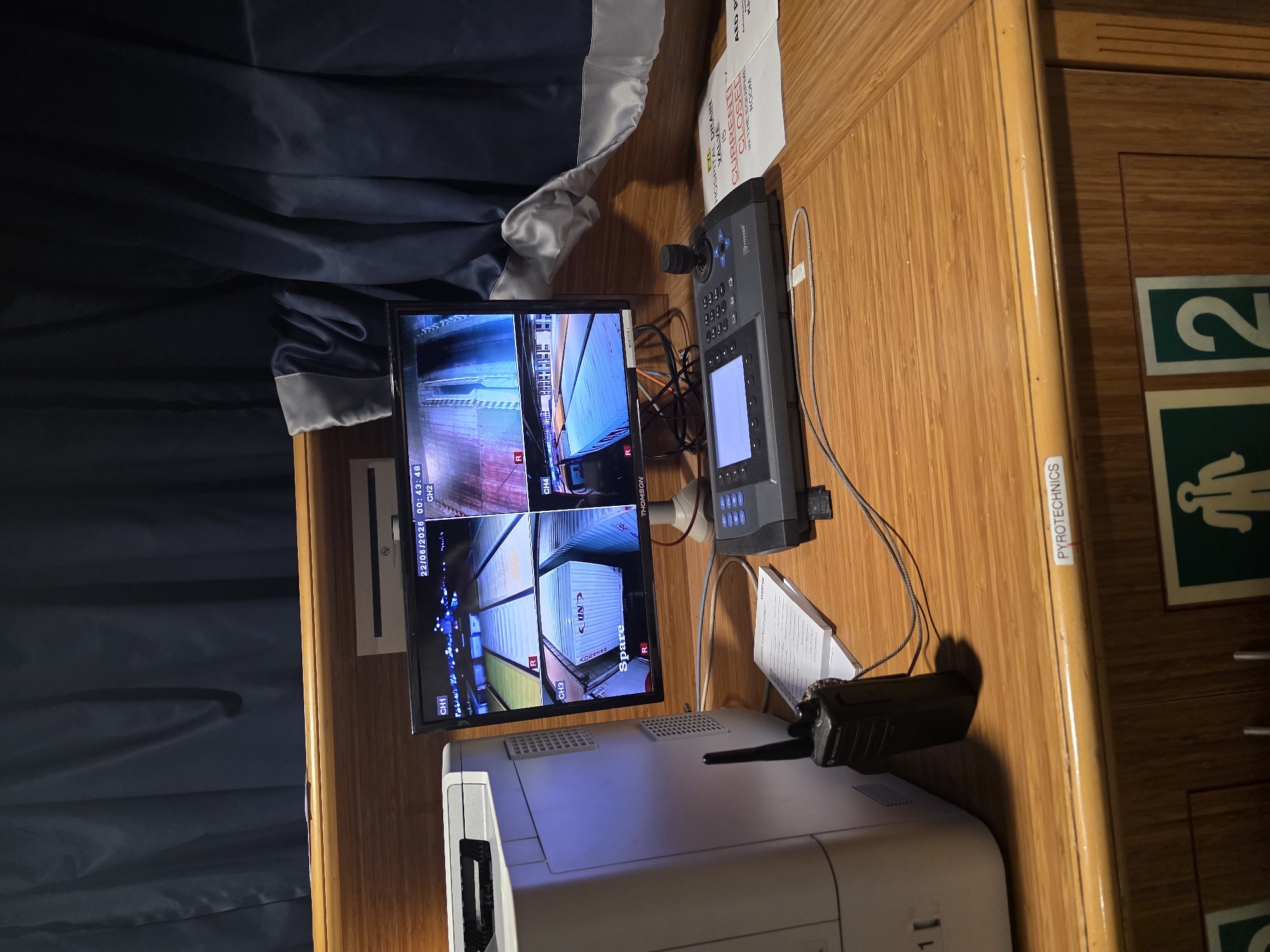 This is a photograph of a security monitoring station likely located in the city of Brindisi, Italy, possibly within a port or industrial facility given the context clues. The scene depicts a wooden desk surface with various electronic equipment.
On the left, a large, off-white Brother printer is partially visible. To its right, a black Motorola radio is positioned on the desk.
Dominating the center of the image is a multi-channel monitor displaying live feeds from four security cameras. The top left shows an overhead view of what appears to be a container yard with brightly lit containers. The top right camera feed shows a similar scene, possibly at a different angle or location, with more illumination. The bottom left camera displays a close-up of a white shipping container labeled "SPARE" and "R". The bottom right camera feed is partially obscured but appears to show a vehicle. The monitor also displays the date "22/06/2026" and time "00:43:48".
In front of the monitor is a control unit with a joystick, a touchscreen display, and a keypad, likely used for operating and navigating the camera feeds. A document, possibly an instruction manual or logbook, is placed beneath the radio.
To the right of the control unit, a sign is visible with red text stating "CURRENTLY CLOSED" and "HOSPITAL MAIN VALUE IS IN THE EQUIPMENT ROOM". Another sign with a green icon depicting a person walking and the number "2" is also visible towards the bottom right. A label on the desk's edge reads "PYROTECHNICS".
The background is dominated by dark blue curtains, which appear to be draped, suggesting an enclosed space like a control room or office. The overall environment appears to be functional and focused on security operations.