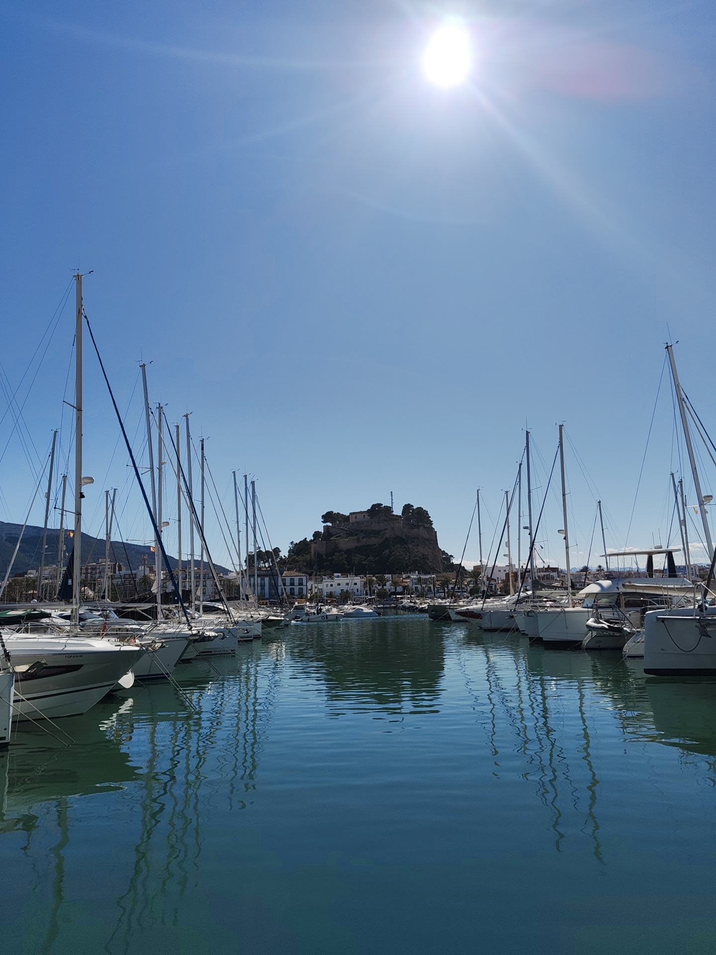 The image depicts a marina in Dénia, Spain, filled with numerous sailboats and motorboats. These vessels are docked along the sides of the marina, their masts creating a dense vertical line. The water is calm and reflects the masts and the boats, causing distorted vertical lines. In the background, a hill is topped by a castle or fortress. The sky is a clear blue, with the sun shining brightly.