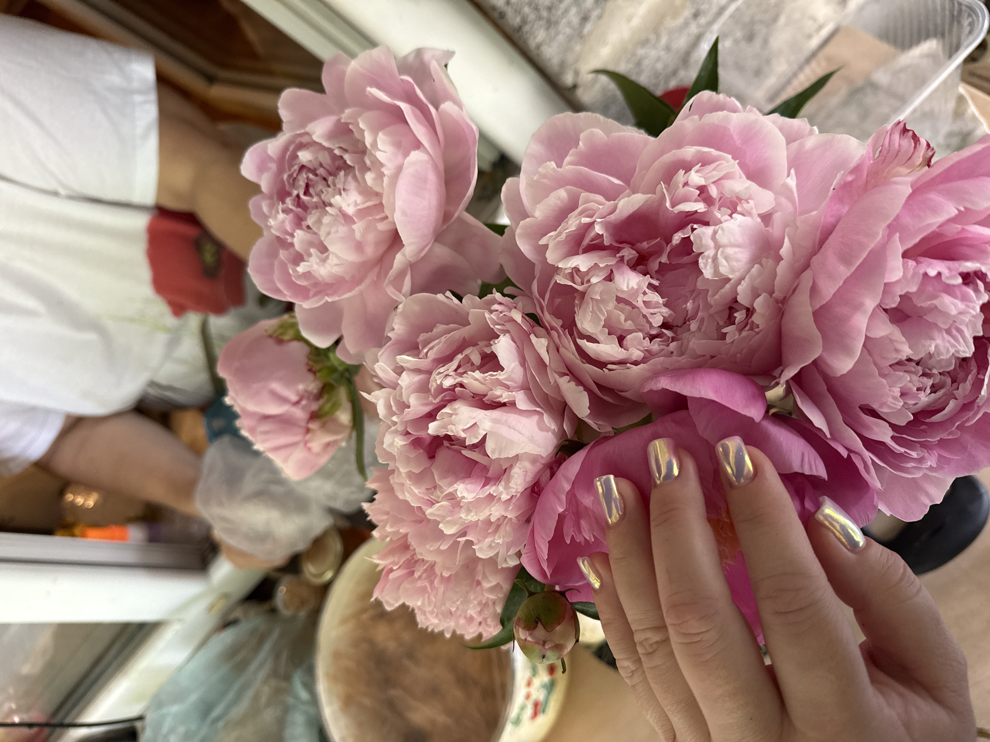 In Nizhne-Chufichevo, Russia, a close-up shot focuses on a bouquet of pink peonies, with a person's hand in the foreground. The hand, adorned with iridescent golden nail polish, gently touches one of the large, layered peony blooms. In the background, partially visible and out of focus, are elements suggesting an indoor setting with diffused lighting, possibly a kitchen or dining area, with a person wearing a white t-shirt in the far background. The overall scene captures a moment of appreciation for the floral arrangement.