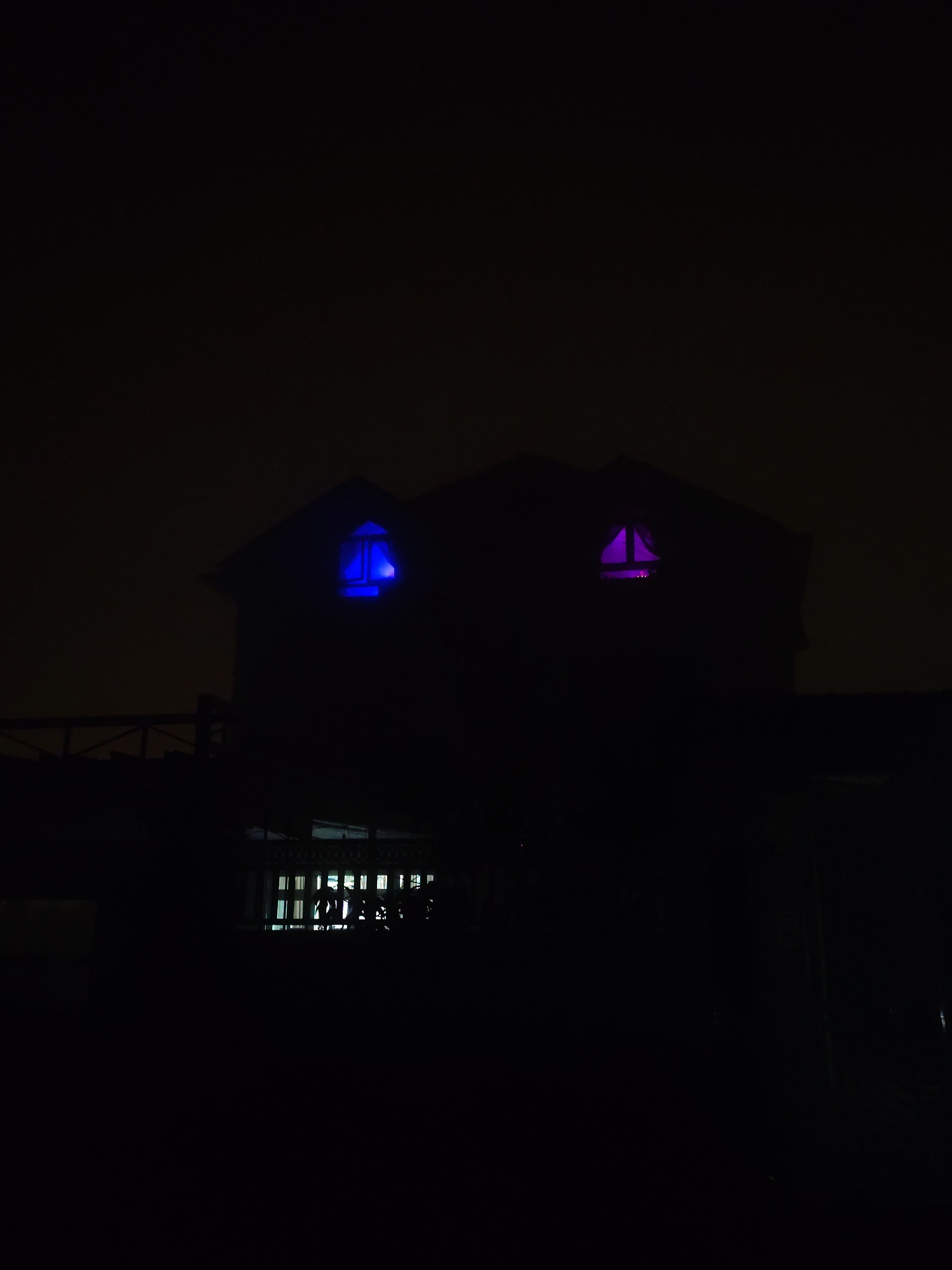 The image presents a nighttime scene of a two-story building or house, silhouetted against a dark, featureless sky in Lạc Dương, Vietnam. The dominant characteristic is the profound darkness of the surroundings, sharply contrasted by artificial light emanating from within the structure.
Two attic windows on the upper level of the building are brightly illuminated with distinct colors. The window on the left glows with an intense, vivid blue light, while the window on the right emits a vibrant magenta or pink light. These colored lights create a striking visual focal point against the otherwise dark facade of the building.
Below the main structure, on a lower level, another window is visible, lit with white light. This window displays a grid-like pattern, likely from a window frame or security bars, and reveals indistinct shapes of objects or foliage inside. To the left of the building, a dark railing or fence structure can be partially seen, blending into the shadows.
The scene is devoid of people or visible activities. The time of day is clearly night, with no discernible weather conditions other than the dark sky. The dramatic interplay of light and shadow, coupled with the unusual colored lights in the windows, imparts a mysterious or perhaps artistic ambiance to the building. No visible text is present in the image.