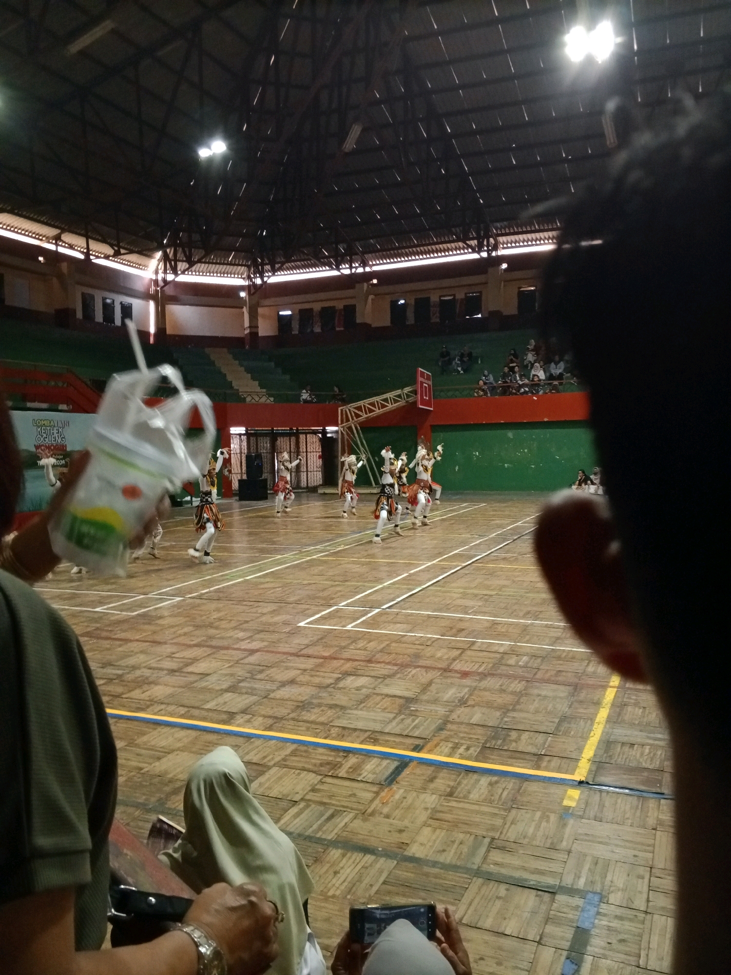 The image shows an indoor sporting arena in Wuryorejo, Indonesia, with a basketball court marked on the wooden floor. A group of performers in traditional costumes are dancing on the court. Spectators are seated in the bleachers. In the foreground, a person is holding a drink and a phone. Another person is partially visible, with their head and shoulder in the frame. The arena has a high ceiling with exposed beams and lights. The walls are painted green and red. The overall lighting is dim, with spotlights illuminating the court.