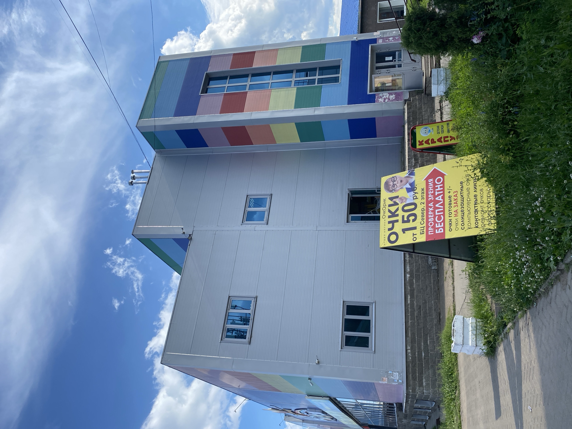This is a photograph taken at street level, with the camera angled upwards, capturing the facade of a modern building and the sky above. The building has a distinctive, colorful design on its upper levels, featuring horizontal bands of rainbow-like colors. Below this, the building transitions to a plain, light-colored metallic siding. Several windows are visible on the facade.
In the foreground, to the right, is a grassy area with a sidewalk made of interlocking paving stones. There are two prominent yellow A-frame signs. The larger sign, in focus, displays text in Russian, advertising an optician's shop. It reads "Очкарик" (Ochkarik) and prominently features "ОЧКИ ОТ 150 руб." (Glasses from 150 rubles). Other text on the sign details services like "ПРОВЕРКА ЗРЕНИЯ БЕСПЛАТНО" (Free vision check) and a list of optical products and services. A smaller sign further back reads "КАРАПУЗЫ" and appears to advertise children's clothing and accessories.
The sky is a bright blue with fluffy white clouds, suggesting a clear, sunny day. The overall impression is a commercial street scene in what appears to be a relatively warm season, given the lush greenery. The building's colorful design hints at a cheerful or family-oriented establishment, possibly a shop or a service center aimed at a broad audience. The presence of overhead wires in the sky indicates an urban or semi-urban setting.