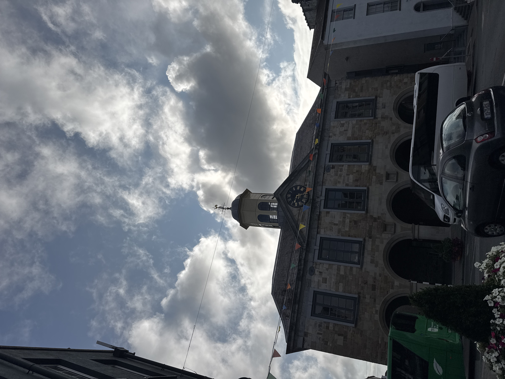 The image shows a low-angle view of the Clonmel Town Hall in Clonmel, Ireland. The building is made of stone with arched walkways on the ground floor and several windows on the upper floors. A clock is visible on the facade of the building, and a weather vane sits atop a cupola. Colorful bunting hangs across the front of the building. In the foreground, a green bus and a white bus are parked, along with a dark-colored car. Some potted plants and flowers are also visible. The sky is partly cloudy with sunlight breaking through.
