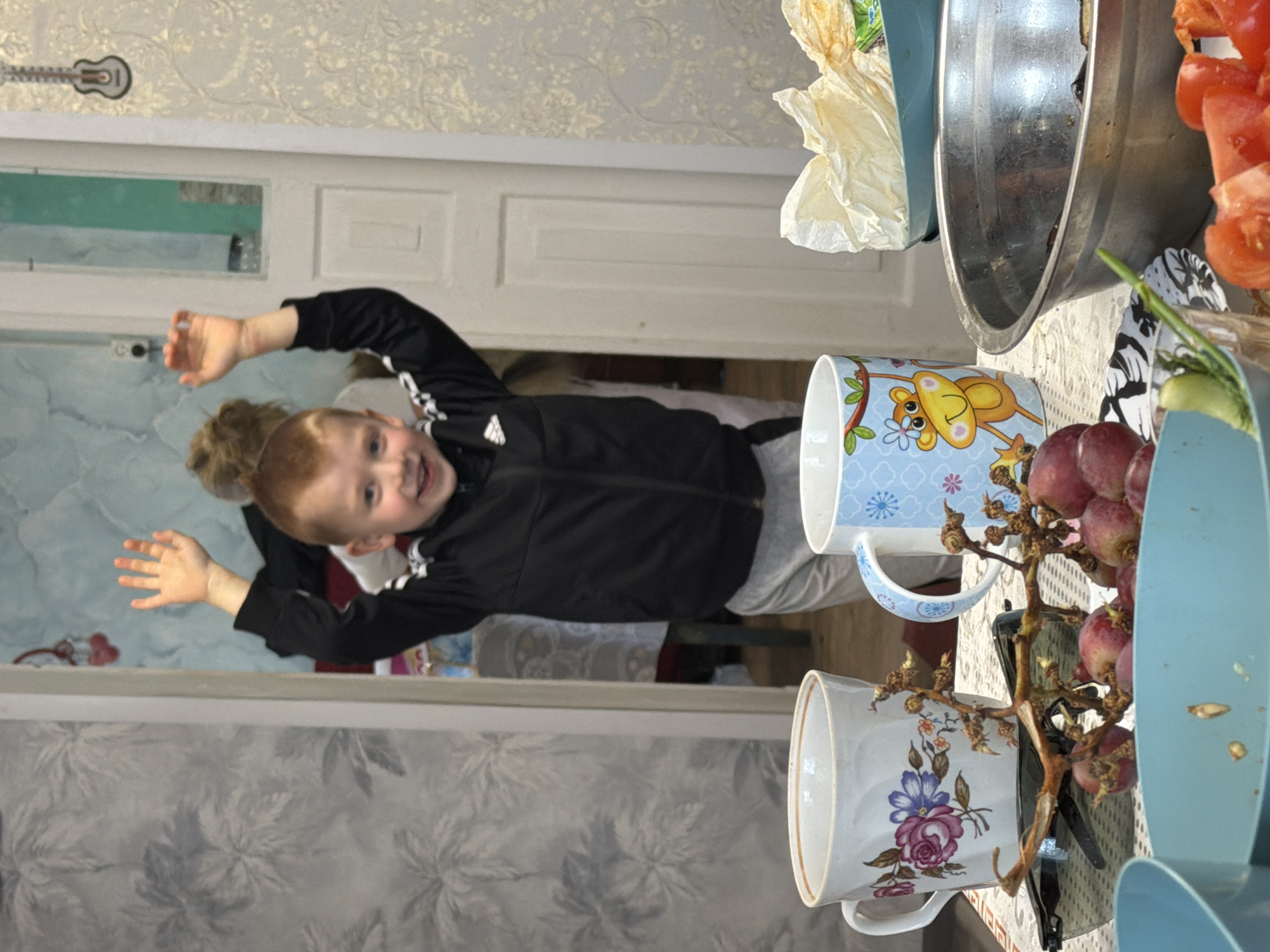 A young boy with a reddish-blonde ponytail is standing in a doorway, facing the camera with arms raised. He is wearing a black Adidas jacket with white stripes on the sleeves and grey pants. The doorway leads into a room with blue floral wallpaper. The wallpaper extends to the left of the boy. To the right, on a table, are two ceramic mugs, one with a blue background and a cartoon monkey, and the other white with a floral design. There is also a bunch of red grapes, a blue bowl, and a large metal bowl with tomatoes and some kind of leafy green inside. The location is Бронница, Russia.