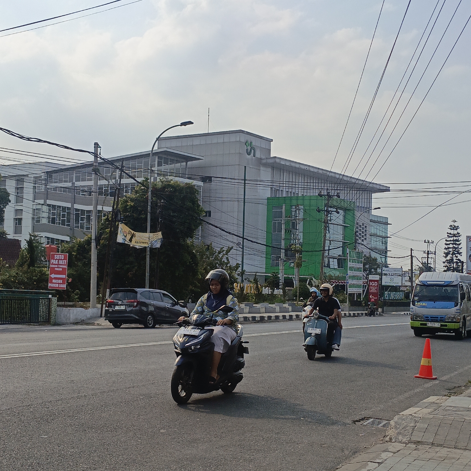The image depicts a street scene in Salatiga, Indonesia, featuring a large modern building with a green accent on its facade, possibly a hospital or a commercial complex. Several vehicles are present on the asphalt road. In the foreground, a woman is riding a dark-colored scooter, wearing a patterned headscarf and grey pants. Behind her, a man is riding a light blue scooter with another person as a passenger, both wearing helmets. Further down the road, a silver car is parked on the side, and a light blue and white van is driving. An orange traffic cone with a yellow stripe is placed on the side of the road. Several utility poles with numerous wires crisscross the sky, indicating an urban environment. Signage is visible, including a red sign with white text indicating "SOTO PAK IKET" and directions for parking, and a yellow banner with text. The sky is overcast, suggesting a daytime scene. The overall impression is a busy street in an Indonesian city with active traffic.