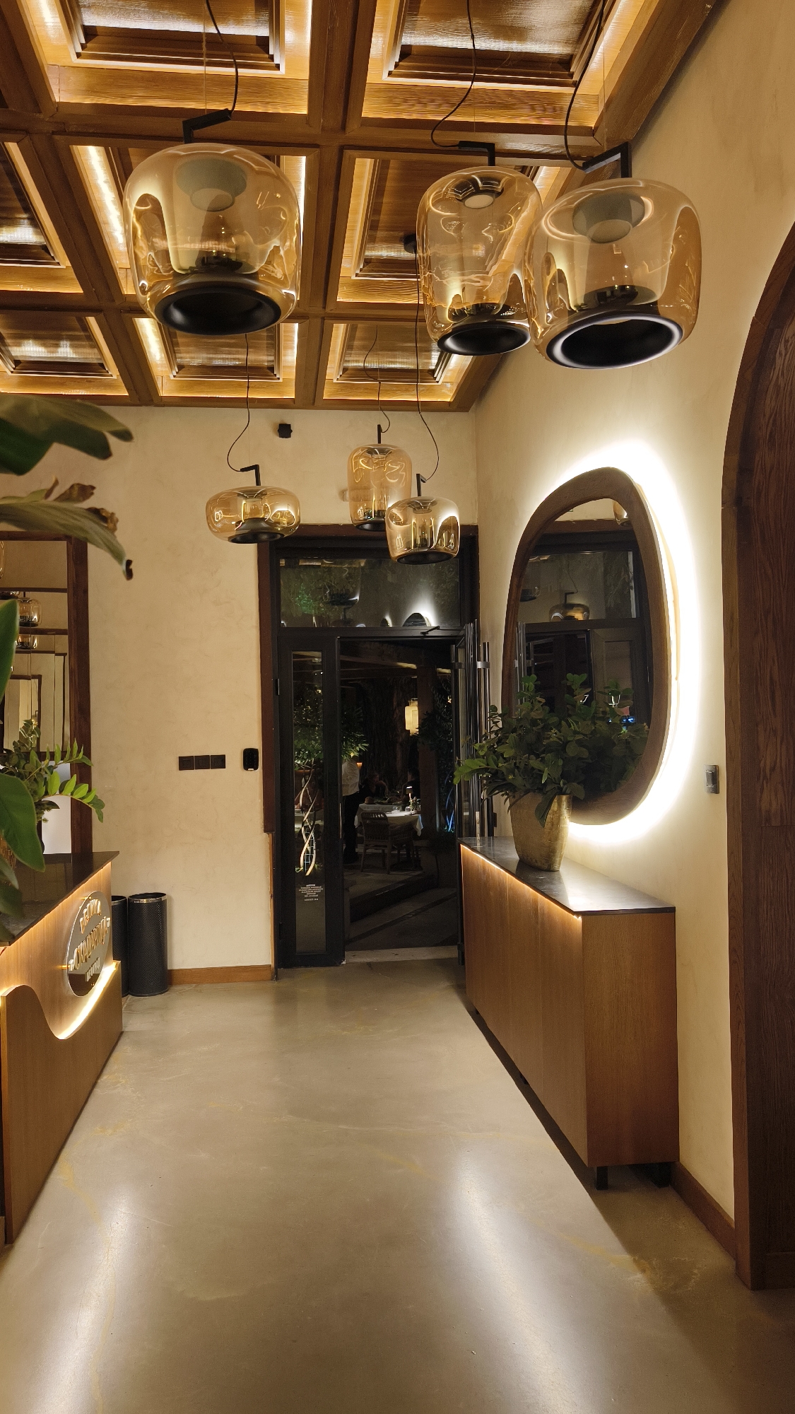 The image depicts a well-lit, modern interior space, possibly a restaurant or lounge, with a contemporary design aesthetic. The ceiling features a grid of wooden beams with integrated warm lighting, creating a warm and inviting ambiance. Suspended from the ceiling are several amber-colored glass pendant lights, adding to the sophisticated decor.
In the foreground, a polished concrete floor reflects the ambient light. To the left, a curved wooden counter with illuminated signage is visible, suggesting a reception or bar area. A black waste bin is placed next to the counter.
The focal point of the midground is a set of double glass doors leading into another area of the establishment. Through the doors, tables and chairs are visible, with some patrons seated. The lighting in this inner area appears to be a combination of overhead fixtures and table lamps, creating a more intimate atmosphere.
To the right of the entrance, an oval mirror with a wooden frame is mounted on the wall, and it is illuminated by a soft, halo-like light emanating from behind it. Below the mirror, a long, wooden cabinet serves as a display surface for a potted green plant in a golden vase.
The walls are painted a neutral, off-white color, and the overall environment suggests a contemporary and upscale venue. The scene is devoid of any explicit markers indicating the specific city or country.