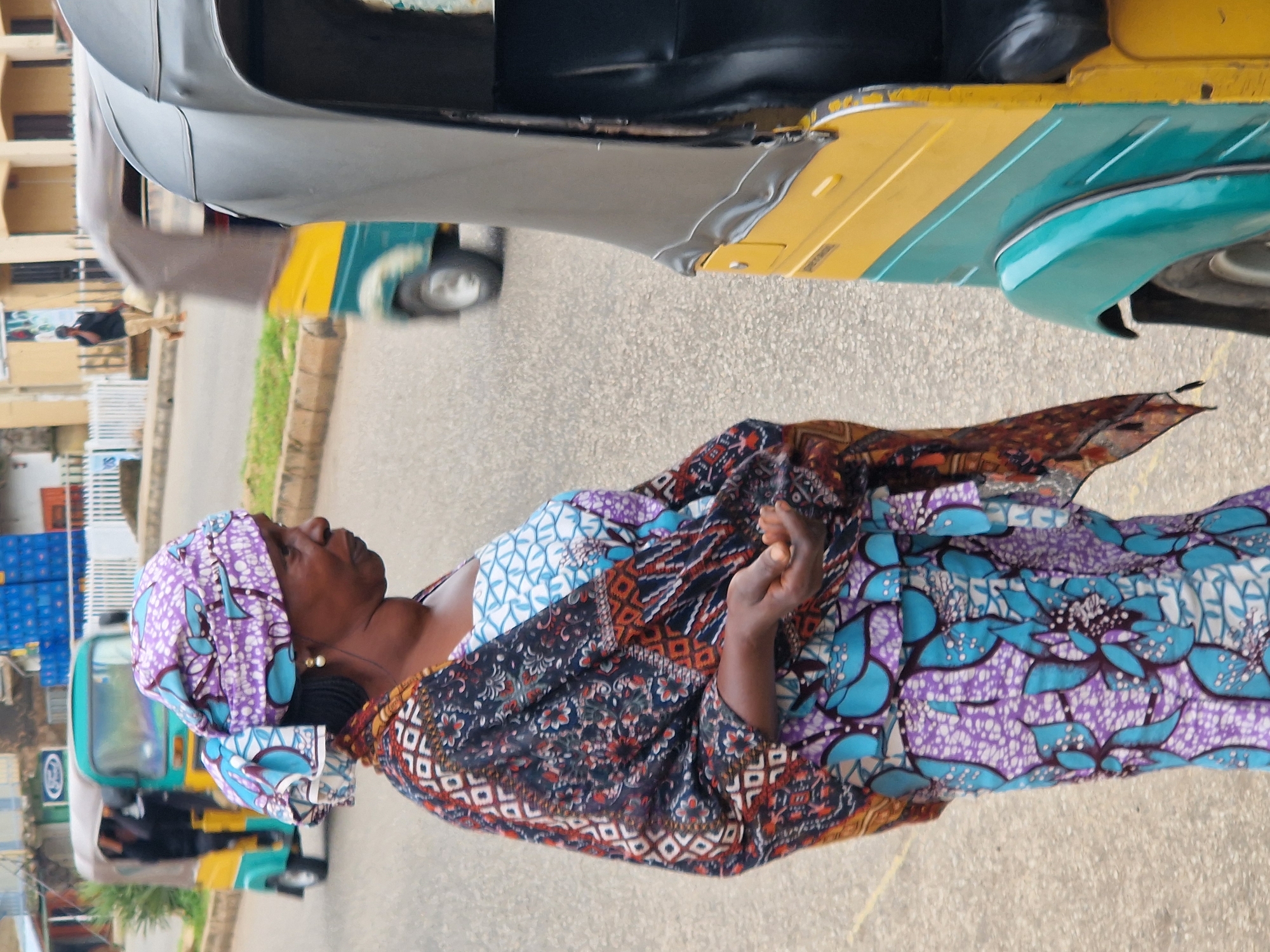 In Kabong, Nigeria, an adult female is captured in a medium shot, standing on a paved road. She is wearing a traditional wrap dress and headscarf, both adorned with vibrant purple and blue floral patterns. A patterned scarf, featuring floral and geometric designs in dark blue, red, and orange, is draped over her shoulders. Her right hand is open, held out in front of her. Behind her, a yellow and teal motorized rickshaw is visible, and further in the background, another rickshaw, primarily green and yellow, is in motion, with a person seated inside. The environment includes a sidewalk with a white fence and a building with a light-colored facade.