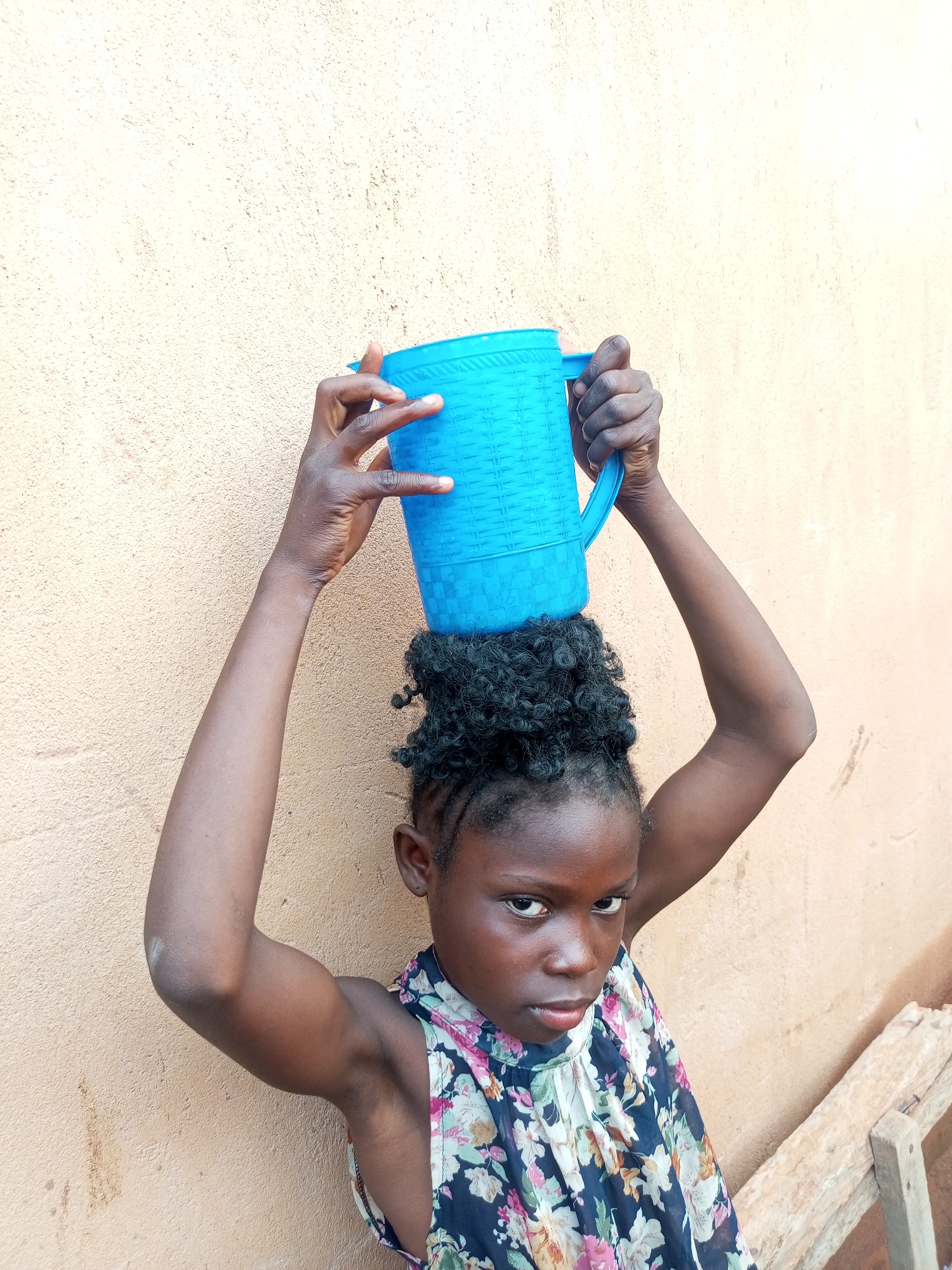 A young Black girl with dark skin, braided hair, and a floral-patterned top balances a bright blue plastic jug on her head. She stands with her arms raised, holding the jug in place. The background is a textured, light-colored wall. The scene appears to be outdoors, possibly in a courtyard or alleyway, with a wooden structure visible on the right side of the frame. The girl is looking directly at the camera with a serious expression. The image does not provide any specific indicators of the city of Tempure, Nigeria, or any other specific geographical location.