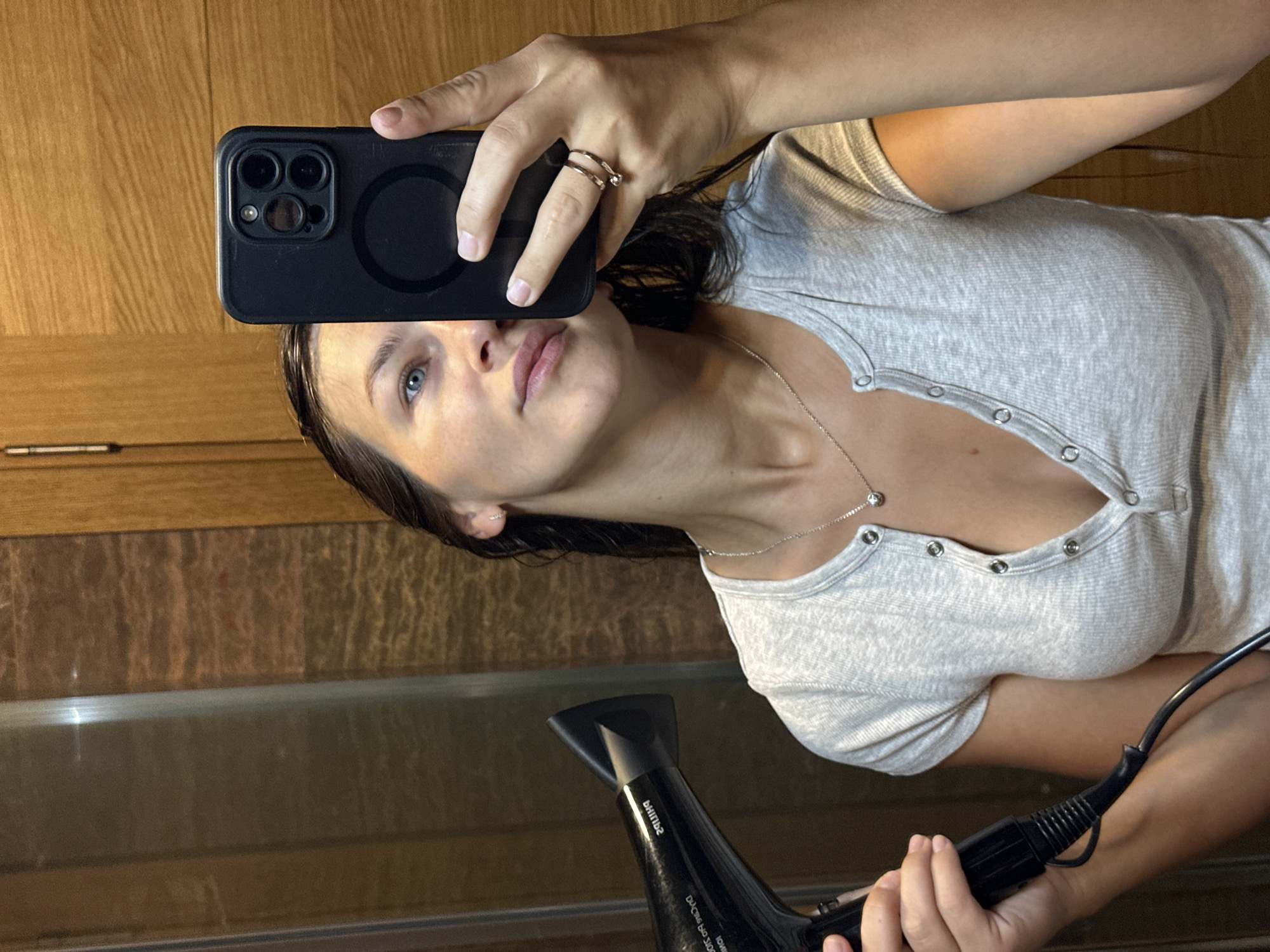 A young woman with wet hair is taking a selfie in a mirror, holding a black smartphone in her right hand and a black hairdryer in her left. She is wearing a light gray, short-sleeved, button-up top. The background appears to be a bathroom with wooden paneling. The scene is captured from a medium shot, focusing on the woman's upper body and face.