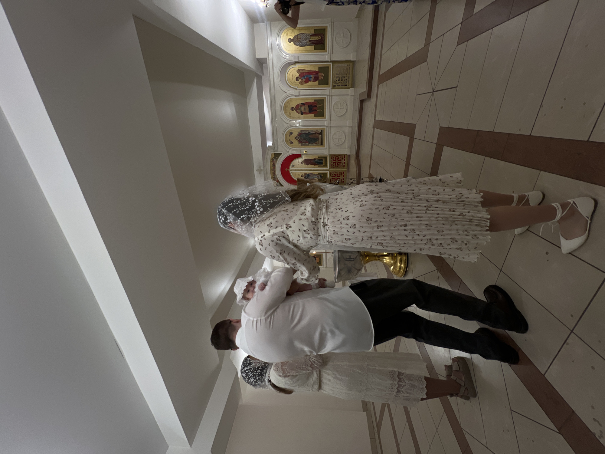 The scene depicts an Orthodox Christian baptism ceremony inside the Church of the New Martyrs and Confessors of Russia, located in Khimki, Russia.
In the center, a man wearing a white long-sleeved shirt and dark trousers holds an infant dressed in a white baptismal gown and bonnet. The man is positioned with his back partially to the viewer, facing towards the right side of the frame. To his immediate left, a woman in a long white lace dress and a dark head covering adorned with a white floral pattern stands observing. To the man's right, another woman, dressed in a long white dress with a delicate floral pattern and a sheer white head covering, stands near a prominent, ornate brass baptismal font. Her gaze is directed towards the infant.
In the background, a white iconostasis spans the wall, featuring multiple gilded arched sections that frame several Orthodox icons depicting various saints and religious figures. The central section of the iconostasis includes red decorative elements, particularly on what appear to be the Royal Doors. The walls and ceiling of the church are uniformly white, with visible structural beams and recessed lighting fixtures integrated into the ceiling. The floor is covered with light-toned rectangular tiles interspersed with darker brown geometric lines, creating a distinct pattern. On the far right, a person, partially visible, holds a black camera, indicating photographic documentation of the event.