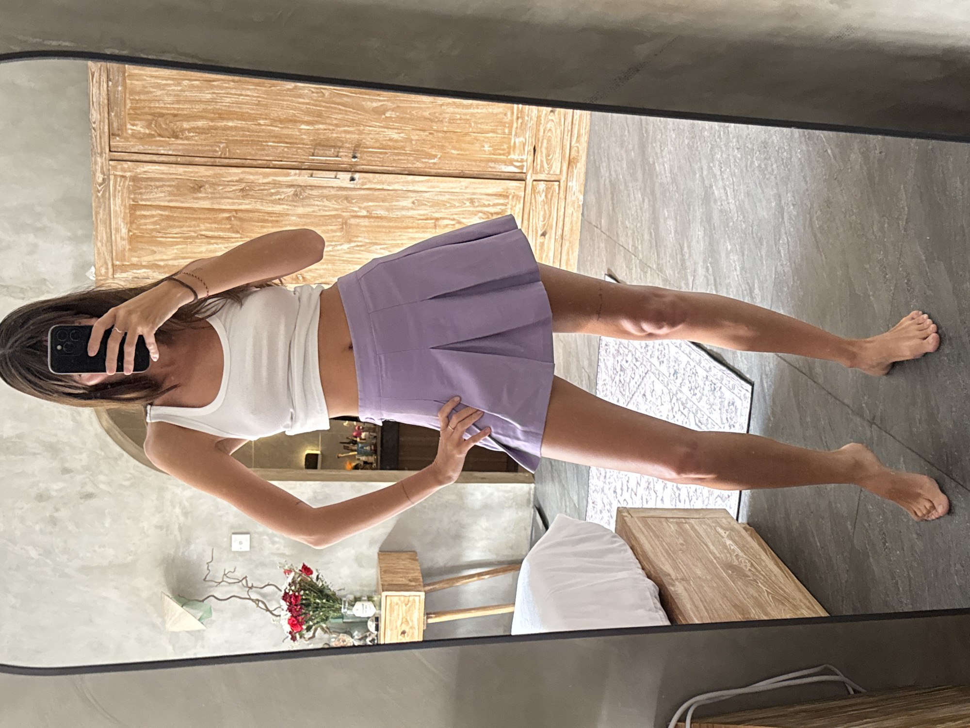 The image shows a woman standing in front of a mirror in what appears to be a bedroom. The woman is holding a smartphone up to take a selfie. She is wearing a white tank top and a light purple pleated mini skirt. The room contains a wooden cabinet, a bed with white linens, and a rug. There are also plants in a vase in the room. The scene is possibly located in North Kuta, Indonesia.