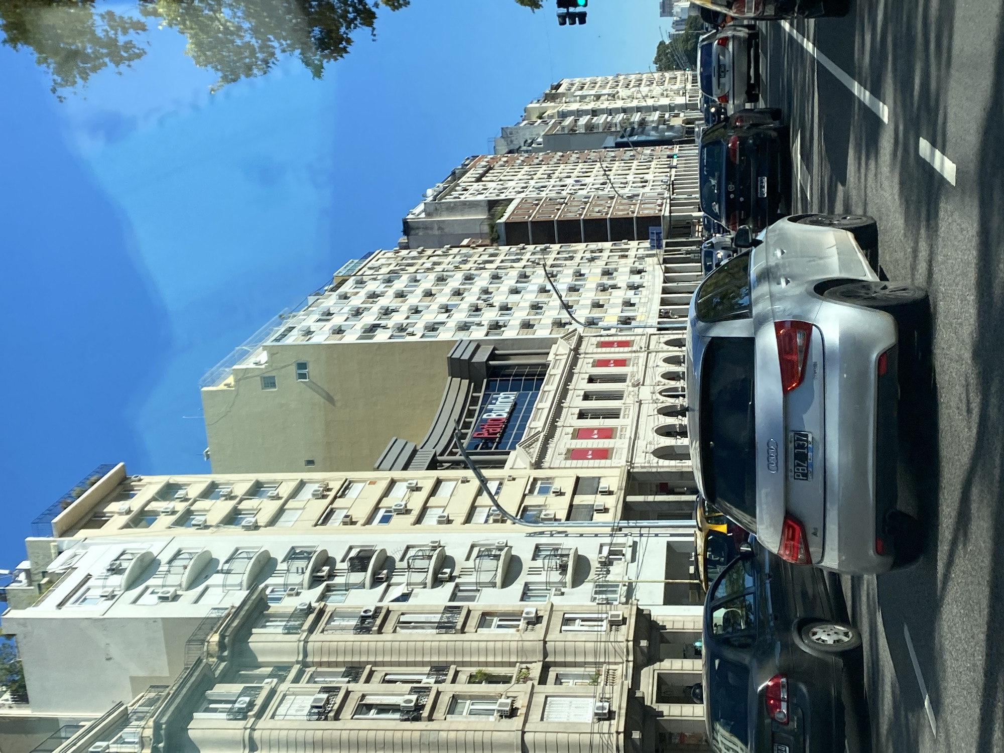 The image captures a street scene in Buenos Aires, Argentina. The camera is likely positioned inside a moving vehicle, offering a perspective of the urban landscape. Several cars are visible, suggesting traffic. Tall buildings line the street, characterized by multiple windows and air conditioning units, hinting at residential or commercial usage. A sign for "Patio Bullrich" is visible on one building, indicating the presence of the Patio Bullrich shopping mall. The sky is clear and blue, indicating good weather. There are no people visible in the image. A traffic light indicates that the light is green.