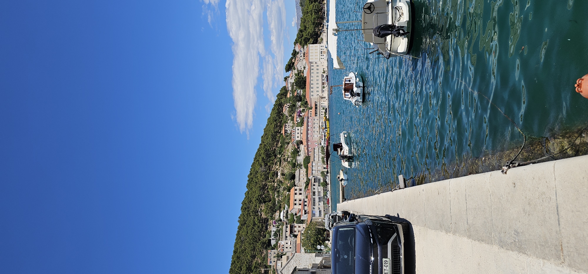 The image depicts a coastal scene in Općina Pučišća, Croatia, under a clear blue sky with scattered clouds. In the foreground, a black car is parked alongside a concrete walkway that borders the blue-green sea. Several small boats are moored in the water, some with canopies. Across the water, a town is built into a hillside, characterized by buildings with reddish-orange tiled roofs, interspersed with green trees and vegetation. The architecture suggests a Mediterranean style. The overall impression is a serene, sunny day in a picturesque seaside town.
