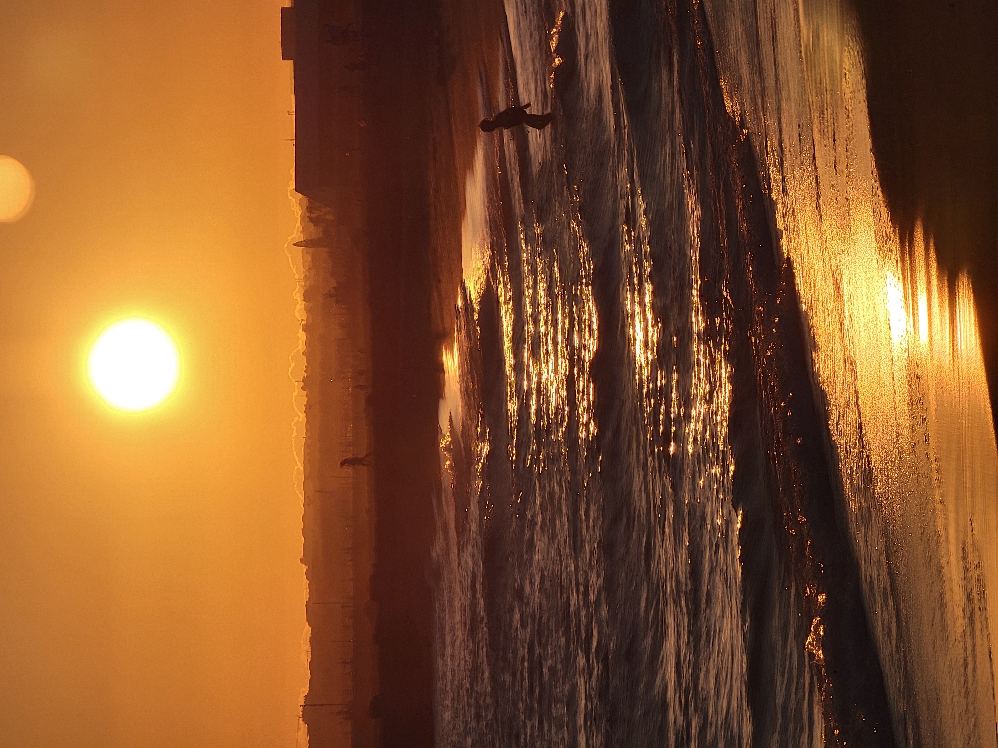 The image depicts a sunset over the ocean at Praia de Quarteira in Quarteira, Portugal. The sky is a vibrant orange, with two bright suns visible. In the foreground, the ocean waves are illuminated by the setting sun, creating a shimmering effect on the water and the wet sand. A lone figure, silhouetted against the light, stands in the shallow water. In the background, the outline of the city of Quarteira can be seen, with buildings and structures dimly visible against the horizon. The overall scene is one of tranquility and natural beauty, with the warm hues of the sunset casting a serene glow over the beach and the sea.