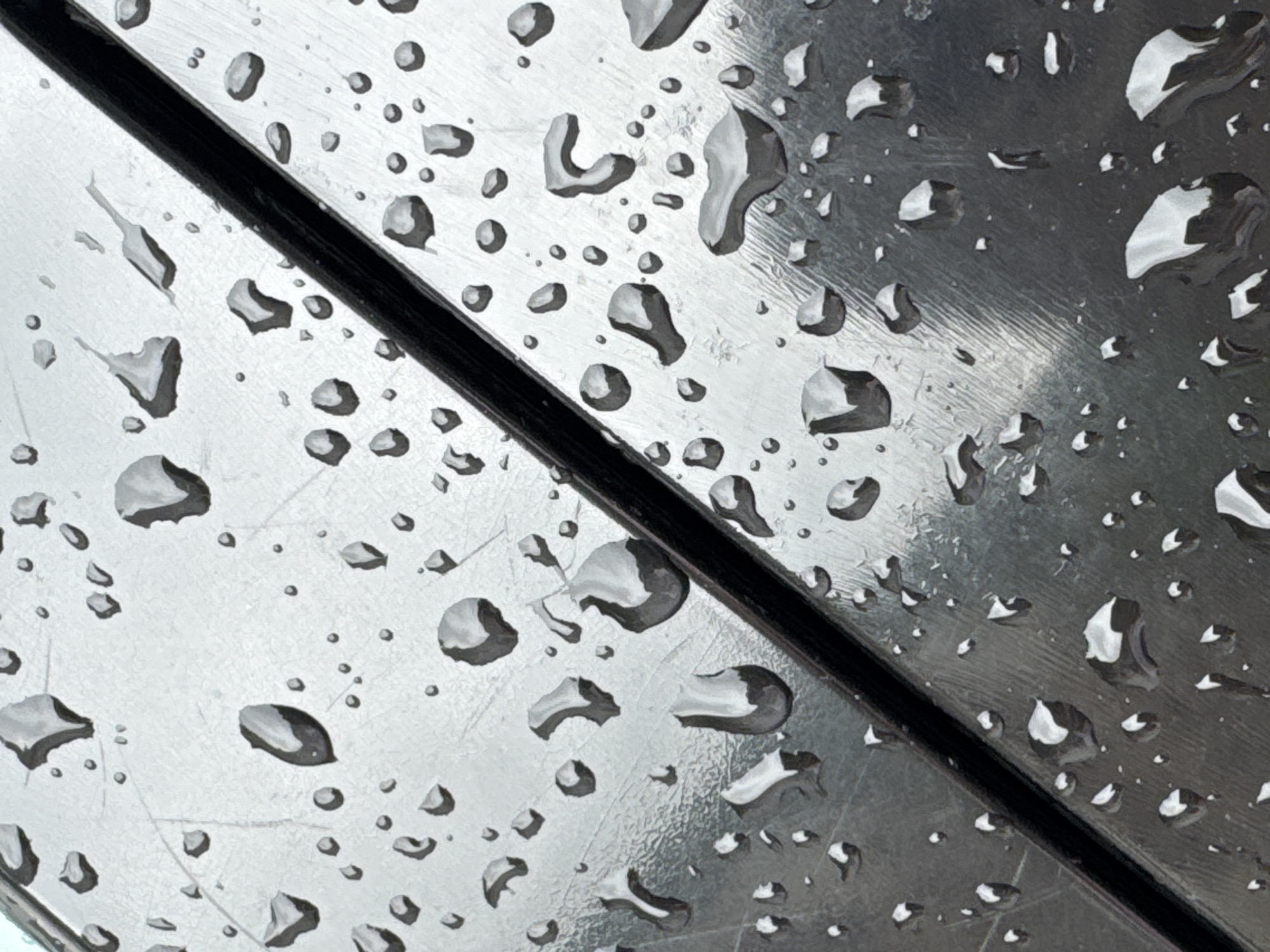 The image shows a close-up of a metallic surface, possibly a part of outdoor furniture like a bench or table, covered in raindrops. A black line or groove diagonally bisects the composition. The surface appears to be reflective, with light creating highlights in the droplets. There's no discernible indication of Budva, Montenegro. The image focuses solely on the surface texture and the presence of water droplets.