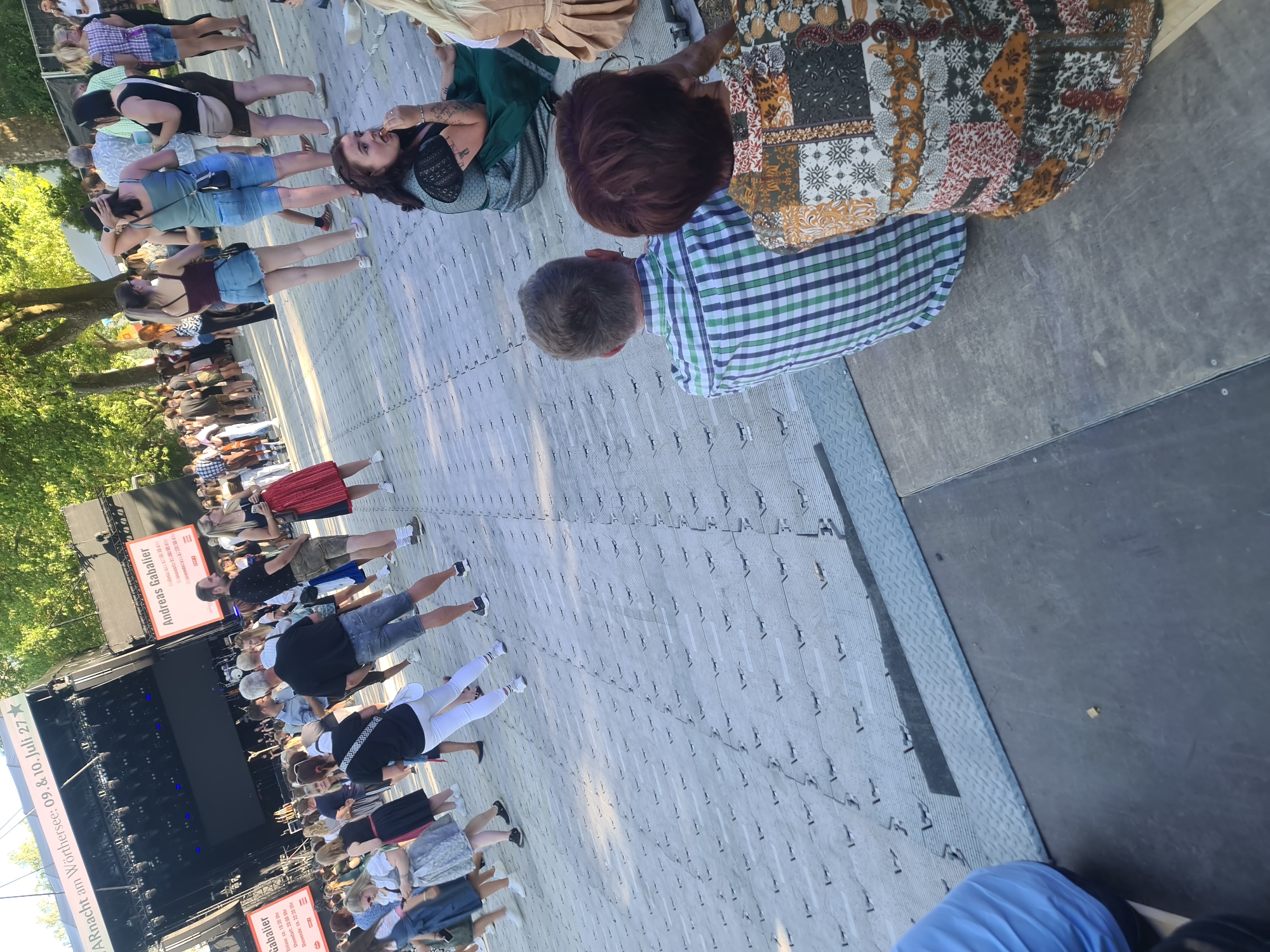 The image captures a sunny outdoor gathering at the Worthersee in Klagenfurt, Austria, likely during a festival or event. In the foreground, two individuals are seated, facing away from the camera. The person on the left wears a blue and green checkered shirt, and the person on the right is adorned in a colorful, patchwork-style top. Behind them, a crowd of people mill about on a patterned ground surface.
The middle ground reveals a large stage, characteristic of an outdoor concert venue, with a black backdrop and various equipment. Several banners are visible in front of the stage, one of which reads "Andreas Gabalier." Signs indicating event dates and times are also present, with text such as "Es lebe die Nacht am Wörthersee: 09. & 10. Juli 27."
The background is dominated by lush green trees, suggesting a park-like setting, and a bright, clear sky. People in the crowd are dressed in a mix of casual attire and traditional Austrian clothing, such as dirndls and lederhosen, hinting at a cultural event. The overall atmosphere appears festive and relaxed, with attendees enjoying the daylight and the event.