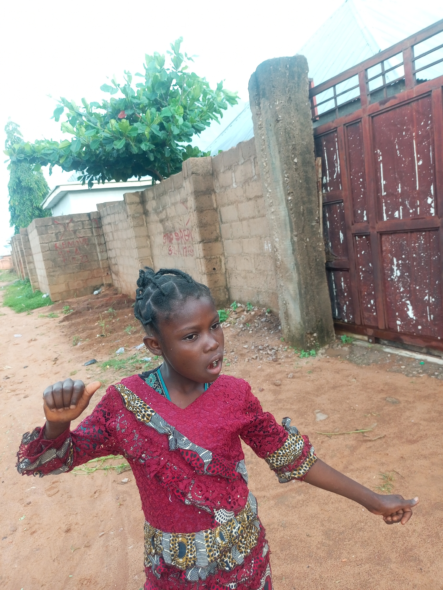 In Tempure, Nigeria, a young Black girl with braided hair is shown in a medium shot, standing outdoors on a dirt path. She is wearing a dark red, lacy dress with colorful patterned accents on the sleeves and waist. Her right arm is extended, with her index finger pointing forward, and her left arm is bent at the elbow, with her fist clenched and thumb extended upwards. Her mouth is open as if she is speaking or singing. Behind her, a brick wall runs horizontally, with graffiti scribbled on some sections. Beyond the wall, there is a large, rusty red metal gate, a thick concrete pillar, and a tree with green leaves. The sky is overcast and white.