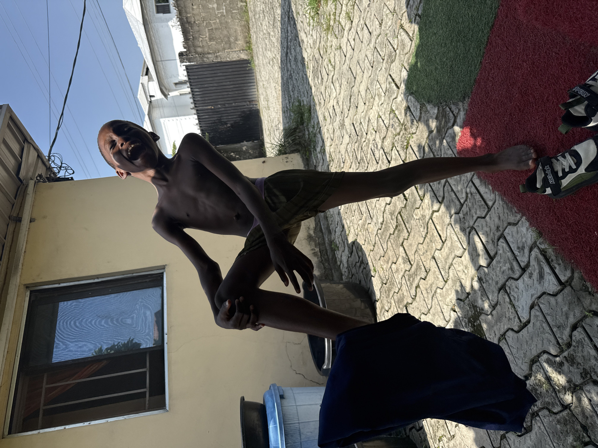 A young boy with dark skin is standing outside in the daytime in Ado, Nigeria, with his mouth open and laughing. He is shirtless and wearing olive-colored shorts. His right leg is bent at the knee, and he is holding his shin with his right hand. A dark blue garment is draped over his left leg, falling towards the ground. The boy is barefoot and standing on a red mat, with a pair of black and white sneakers next to his feet. The ground around him is paved with interlocking concrete blocks, and there is a patch of green turf to the right of the mat. In the background, there is a cream-colored wall with a window, a dark metal gate, and several white buildings with metallic roofs. The sky is clear and blue.
