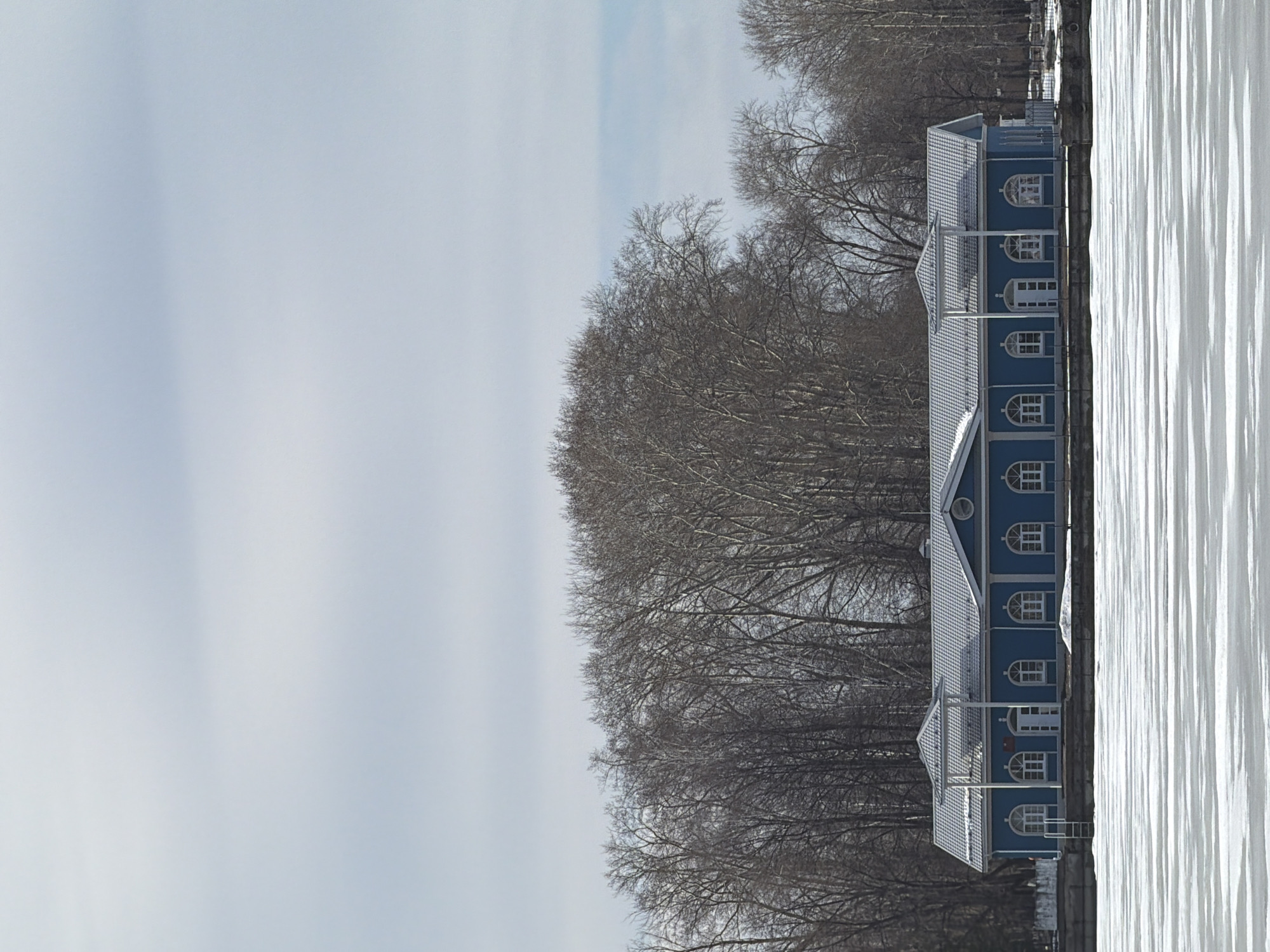The image shows a one-story, light-blue building with a gray roof located on the bank of a snow-covered body of water. The building features a series of arched windows and doors. The building is in Votkinsk, Russia. Bare trees without leaves stand behind the building. The sky is pale and overcast. The body of water is heavily textured, suggesting a layer of ice or compacted snow.