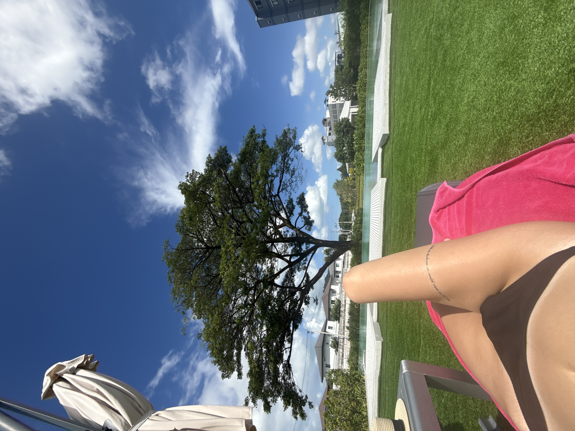 The image is a point-of-view shot from someone relaxing on a lounge chair by a pool on a sunny day in Ratsada, Thailand. The foreground shows a person's legs and torso, wearing a dark bikini bottom and a pink towel draped over their legs. A thin tattoo is visible on the upper thigh. To the left, a portion of a lounge chair and a straw hat are visible. Beyond the lounge chair is a bright green lawn with a large, mature tree casting shadows. In the mid-ground, a swimming pool with white tiling and clear water is present. Behind the pool, houses and buildings are visible, suggesting a residential or resort area. The background features a clear blue sky with scattered white clouds and a tall, dark building on the right side of the frame. The overall scene depicts a leisure setting with a tropical ambiance.