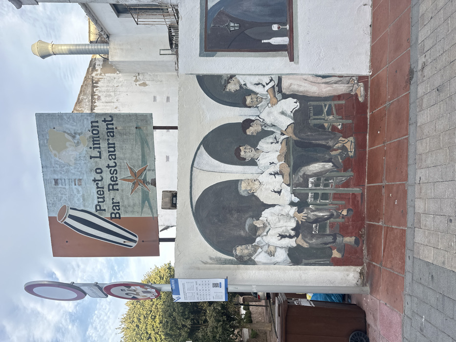 This image depicts a building wall in Calella De Palafrugell, Spain, featuring a large mural and an overhead sign.
Above the wall, a weathered rectangular sign for "Puerto Limón Bar Restaurant" is displayed. It features a striped, elongated drum graphic on an orange panel and a reddish-orange five-pointed star below the text, set against a light blue background.
The wall below the sign hosts a large mural. This mural portrays six men in what appears to be an interior bar or tavern setting with three prominent dark arches. Five men are seated around a dark rectangular table, all wearing white collared shirts and dark trousers; one seated man has a dark flat cap. A sixth man stands to the right of the table, holding a small glass, engaging in conversation with a seated man. The mural's floor is painted in reddish-brown tiles. To the immediate right of the main group, a painted window on the mural shows a wine bottle and a glass containing red wine on a sill, with an interior view of a hanging lantern beyond.
To the left of the mural, a vertical pole stands, bearing three signs: a red circular no-entry sign, a red circular sign with "2T" and "27" possibly indicating a vehicle restriction or towing information, and a white rectangular "TARIFES" sign listing prices. At the base of this pole, on the sidewalk, are two brown wheeled refuse bins and a blue plastic bag.
The ground in the immediate foreground consists of grey paver stones, transitioning to reddish-brown square tiles directly in front of the mural wall. The background above the mural and behind the sign reveals parts of an older, light-colored building, including an exposed metal vent pipe on an upper level and a small balcony with a metal railing. The scene is illuminated by daylight under a clear sky.