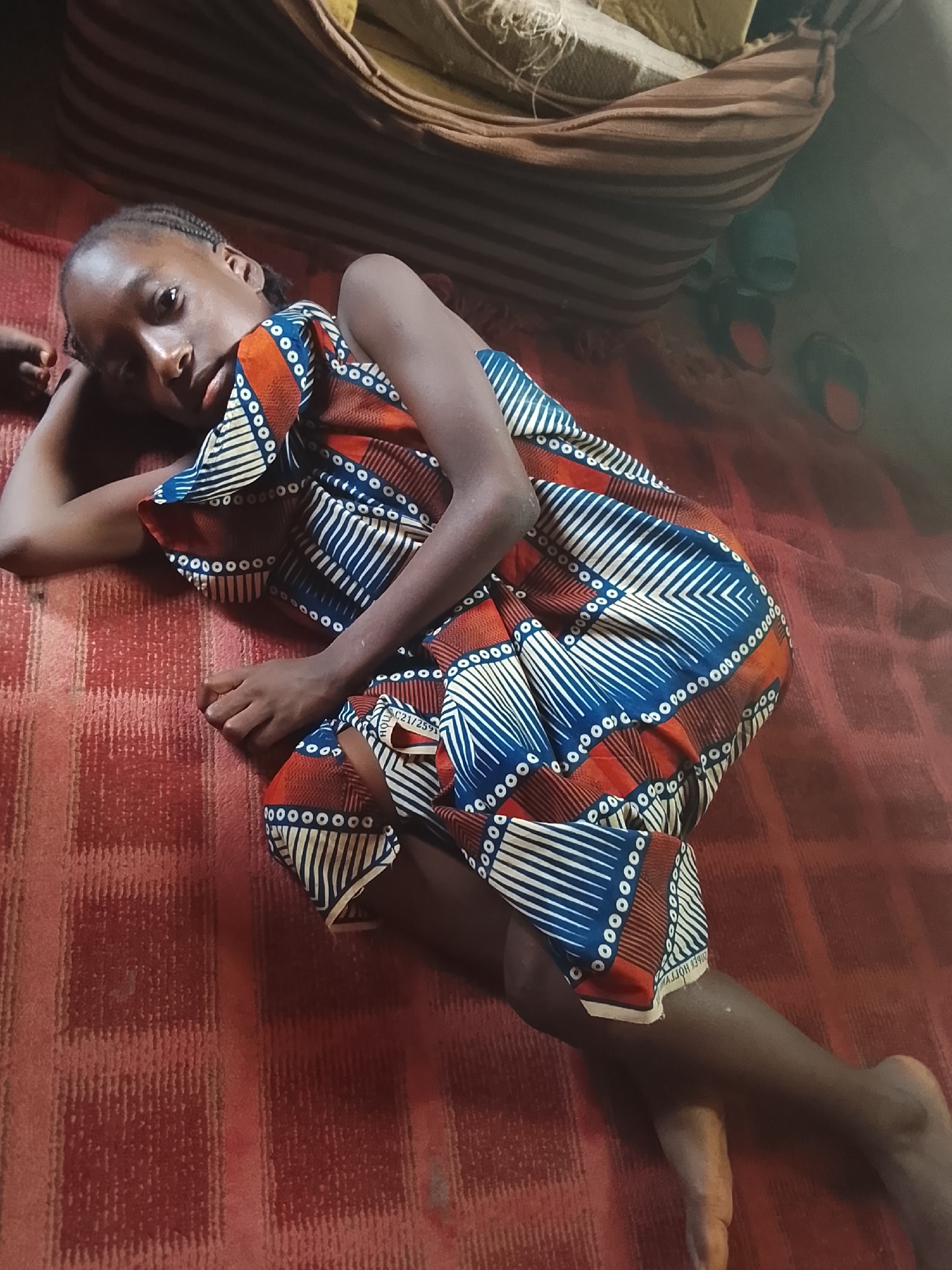 A young person lies on a reddish-brown fabric with a grid-like texture, possibly a blanket or mat. The individual has dark skin and is wearing a brightly colored dress with patterns of blue, white, and orange. Their hair is braided close to the scalp. They are reclining on their side, with their head resting on one hand, and their gaze directed at the viewer.
In the background, there is what appears to be a striped cushion or mattress. A pair of slippers are visible behind the individual, suggesting the setting may be an indoor residential space.