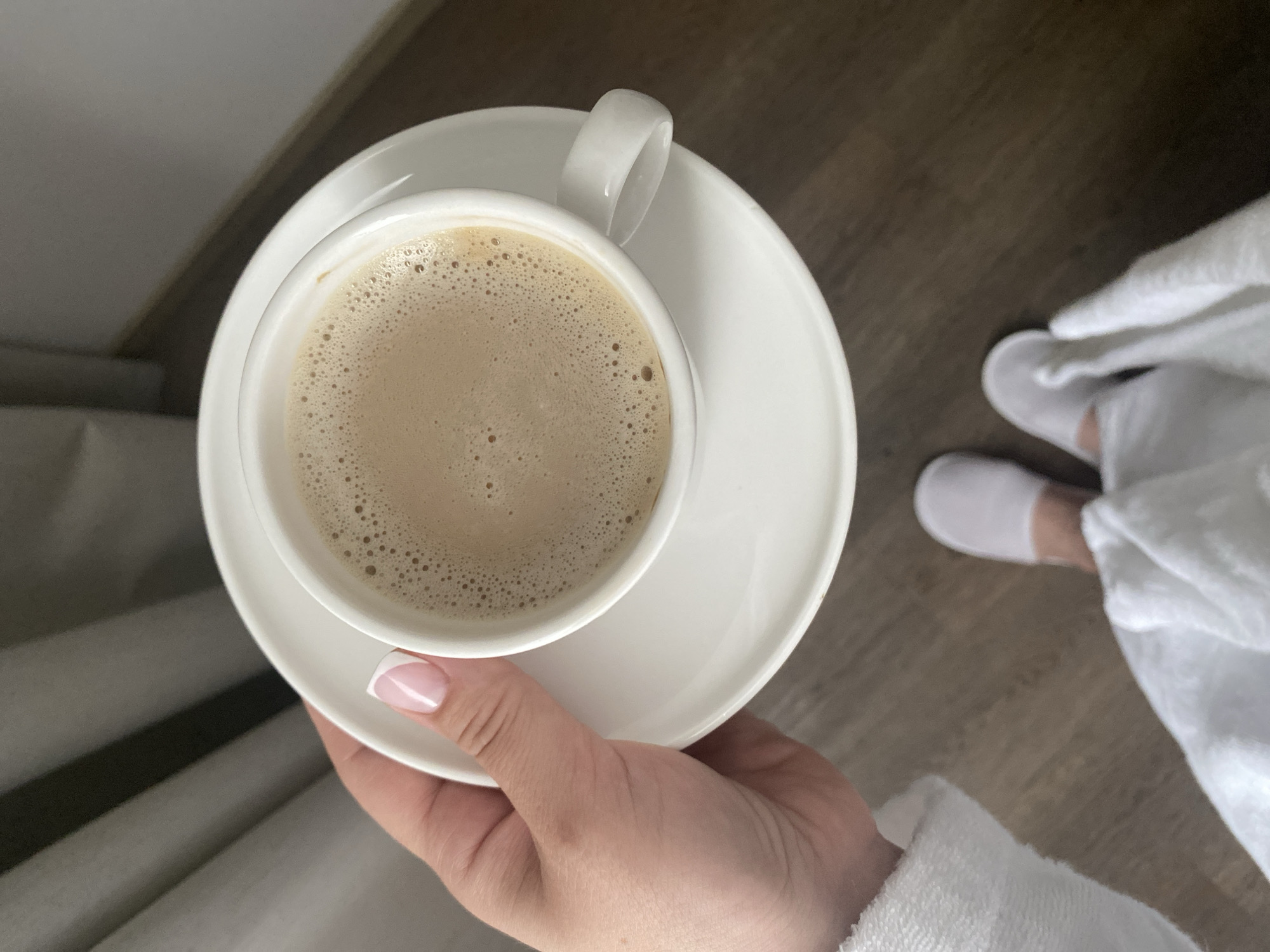 The image depicts a person in a white bathrobe and white slippers holding a white cup and saucer filled with a light brown, foamy beverage. The person's hand has manicured nails with a French tip design. The background shows a wooden floor. There is no information to confirm if the location is Sochi, Russia.