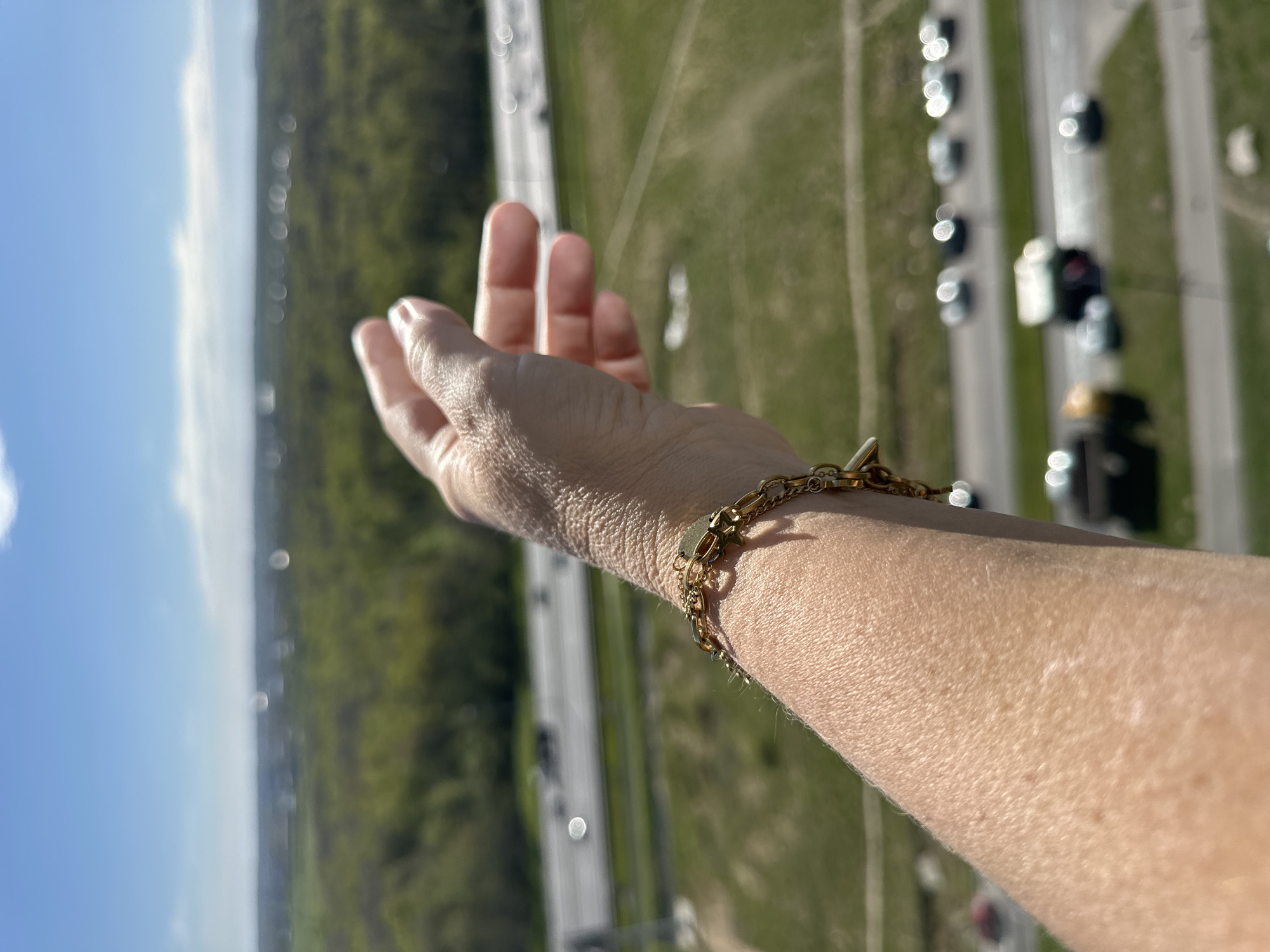 A first-person, aerial view of a human hand and forearm extended outwards, with a gold bracelet visible on the wrist. The background consists of a landscape with a highway running through a green, wooded area and a portion of a clear, blue sky with clouds. Vehicles are present on the highway.