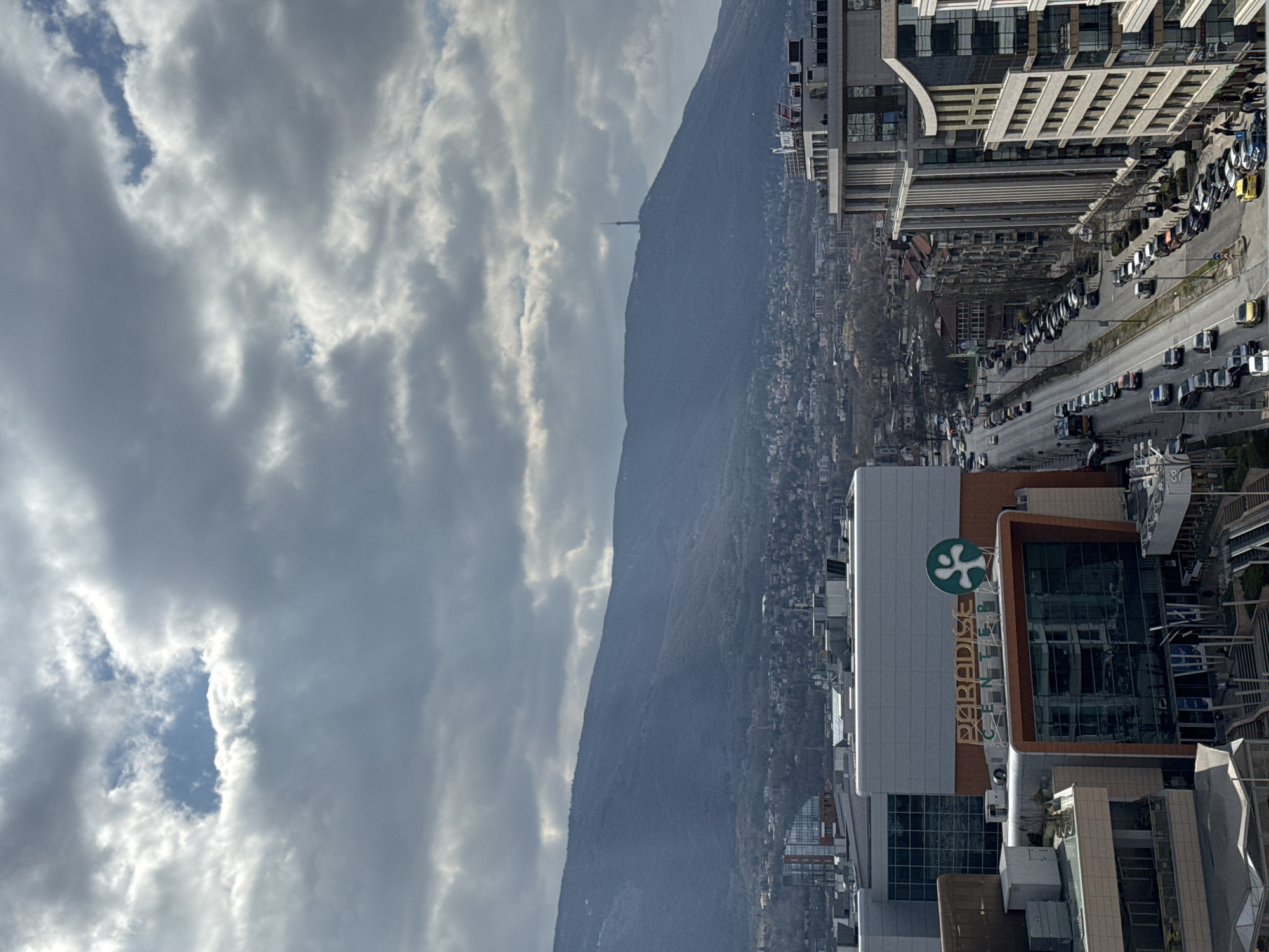 An aerial view of Sofia, Bulgaria, primarily depicting the Paradise Center shopping mall and Cherni Vrah Boulevard. Vitosha Mountain, featuring the Kopitoto TV Tower, dominates the background under a sky largely covered by gray clouds with scattered patches of blue.
In the midground-left, Paradise Center, a large multi-story commercial building, displays its "PARADISE CENTER" signage in gold letters alongside a green, stylized tree logo. The building features extensive glass facades and light-colored paneling. Cherni Vrah Boulevard runs diagonally from the lower center towards the midground, bordering the mall. The boulevard contains multiple traffic lanes and is lined with numerous parked passenger vehicles on both sides. Several vehicles are in motion, including a public transit bus. To the right of the boulevard, a sequence of modern, multi-story commercial and residential buildings with varied facades, including glass and concrete, line the street. Further into the background, a dense urban and suburban residential area extends to the base of Vitosha Mountain. Pedestrians are visible on sidewalks along the boulevard and near the buildings. Deciduous trees throughout the scene appear bare, suggesting a colder season.