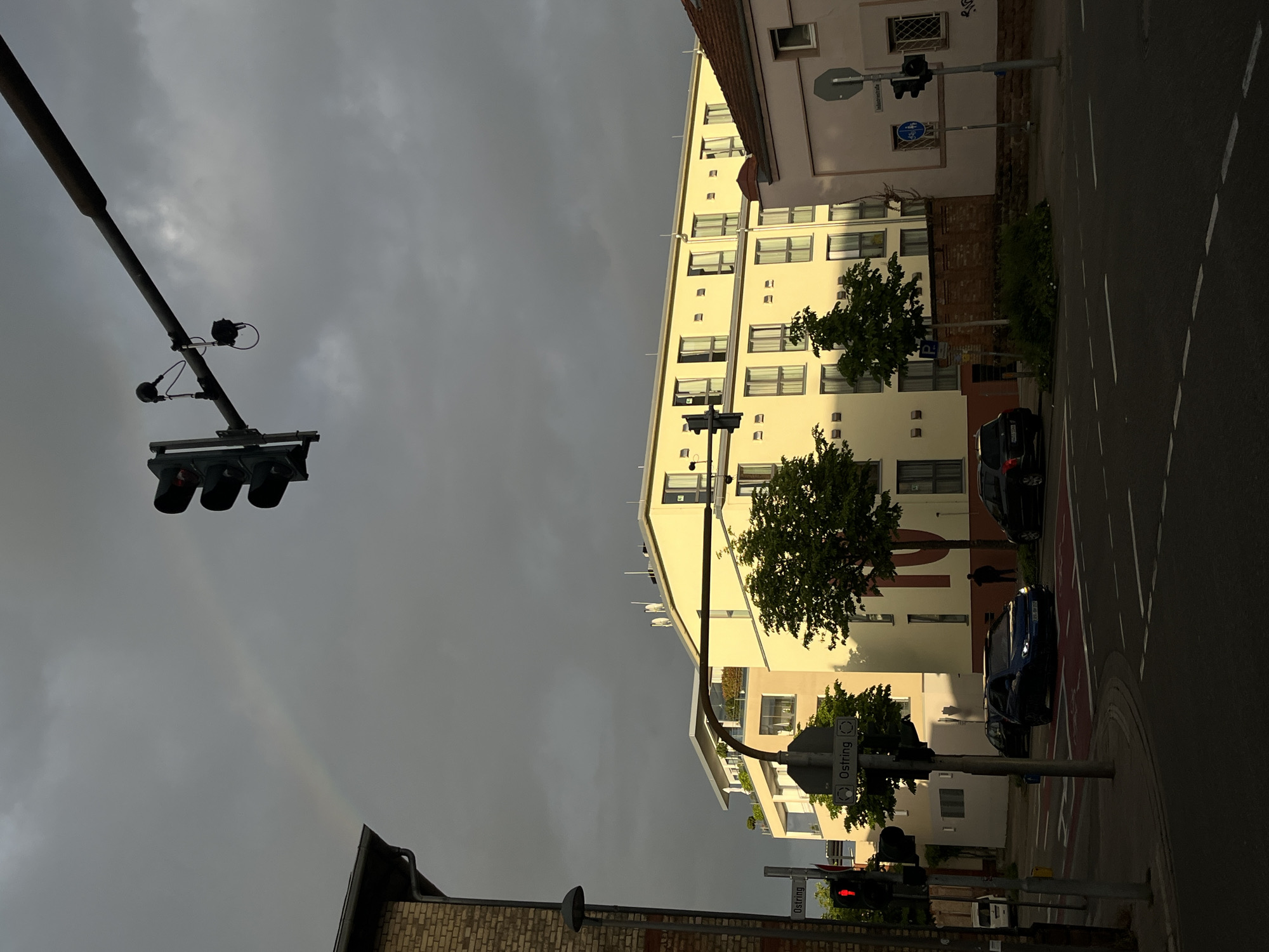 The image captures an urban intersection in Landau in der Pfalz, Germany, under a heavy, overcast sky that exhibits a faint rainbow. A tall, cream-colored building with rows of windows stands along the street, bordered by trees. Several cars, including a blue sedan and a black SUV, are parked or passing by. Traffic lights are visible on both sides of the intersection. Street signs with "Ostring" are present. A tall traffic light pole with signal lights and additional sensor attachments is visible in the foreground. Brick buildings with dark roofs are also visible at the bottom.