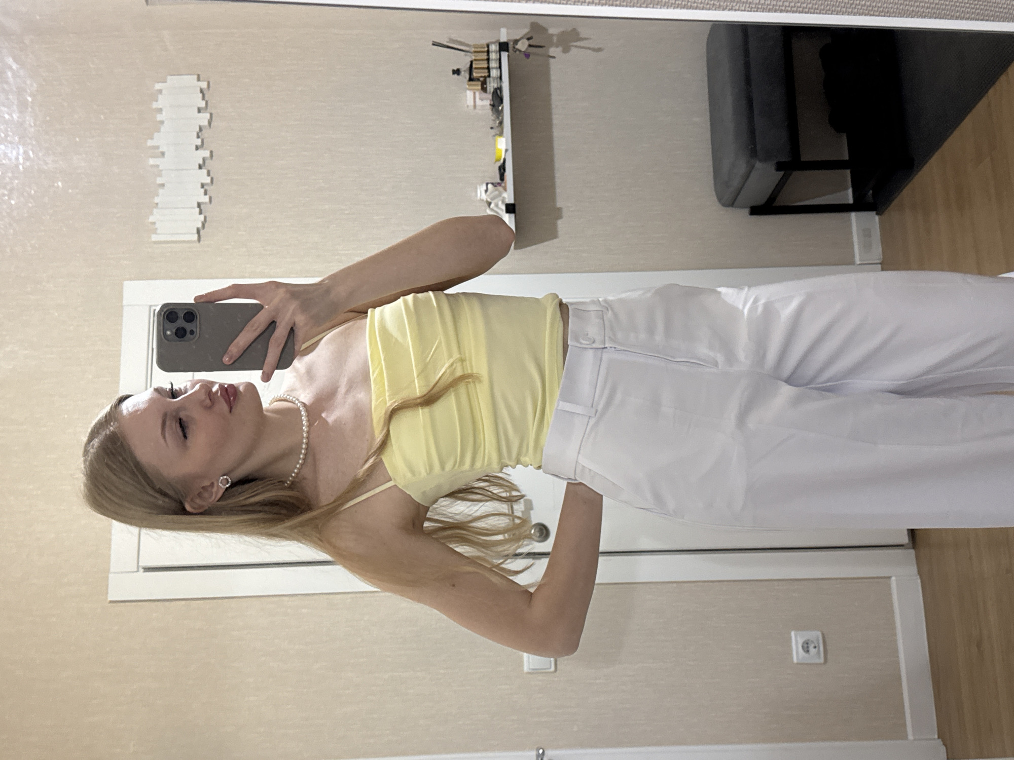 The media file is a portrait of a young woman taking a mirror selfie in what appears to be a bedroom or dressing area. She has blonde hair and is wearing a yellow ruched top, a pearl necklace, and white pants. Her left hand is holding a dark-colored smartphone, capturing her reflection. The background includes a wall with textured wallpaper, a white door frame, a decorative white item mounted on the wall, and a shelf with various items on it. A dark bench or stool is visible in the reflection, along with a wooden floor. The lighting suggests it is daytime indoors. The woman's expression is neutral, and the overall mood is casual and personal. No visible text is present in the image. The location context provided is Perm, Russia.