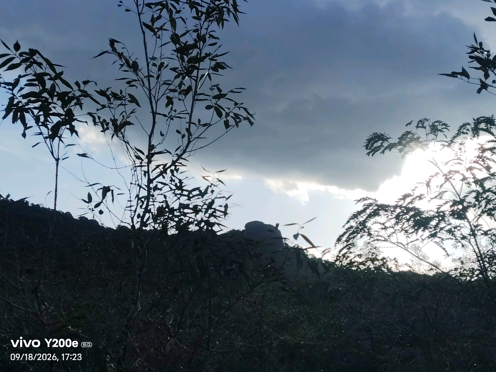The image captures a silhouetted landscape with dramatic cloudy skies, likely taken at the Tippagondanahalli Reservoir in India. In the foreground, dark silhouettes of trees and foliage obscure the ground. A rocky outcrop or hill is visible against the sky in the midground, also silhouetted. The sky is dominated by heavy, dark clouds with areas of bright light breaking through, suggesting either sunrise or sunset or a storm clearing. The bottom left corner of the image displays a "vivo Y200e 5G" watermark and the date "09/18/2026, 17:23". No people or specific actions are discernible.