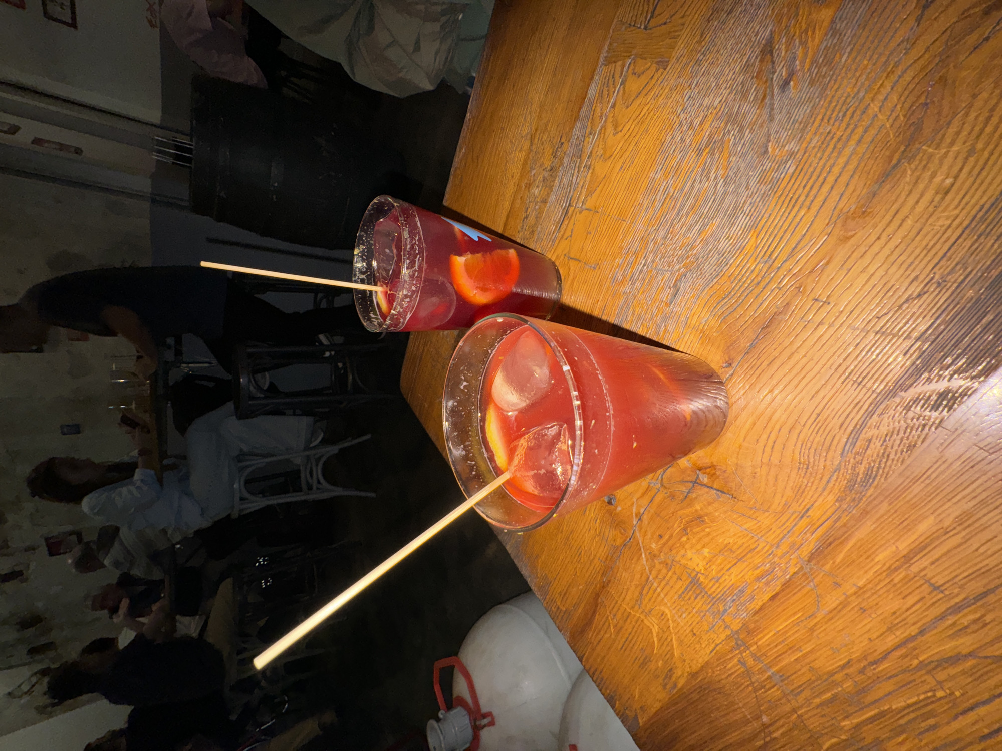 The scene shows two cocktails in tall glasses with long, light-colored sticks or straws, sitting on a heavily textured wooden table. The drinks appear to be red, possibly sangria, with ice and slices of orange. The table exhibits a distinctive wood grain pattern.
In the background, there's a dimly lit area with seated individuals, suggesting a bar or restaurant setting. The location is Barcelona, Spain. Additional details visible include chairs, clothing, and possibly other beverages or items on tables. Some objects near the wooden table base are blurry, including what looks like a pair of white tanks or similar cylindrical objects.