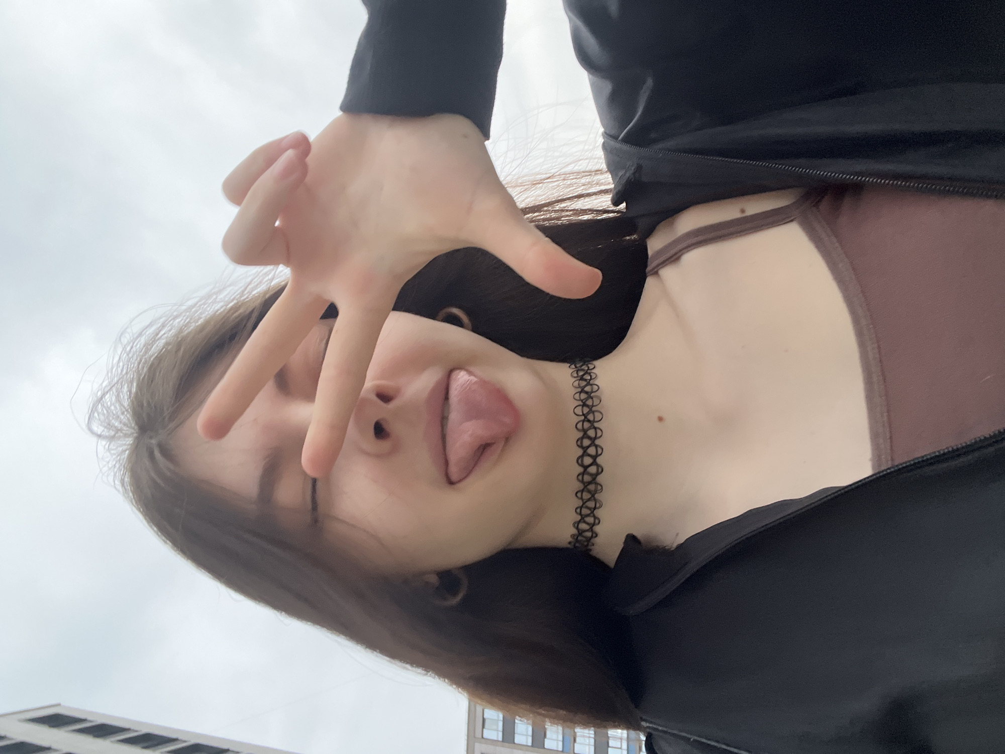 A young woman poses for a selfie outdoors, likely in Saint Petersburg, Russia. She is holding her hand up, obscuring part of her face, and sticking her tongue out playfully. She is wearing a dark jacket over a brown top and a black choker necklace. In the background, a building with multiple windows is visible. The sky is overcast.