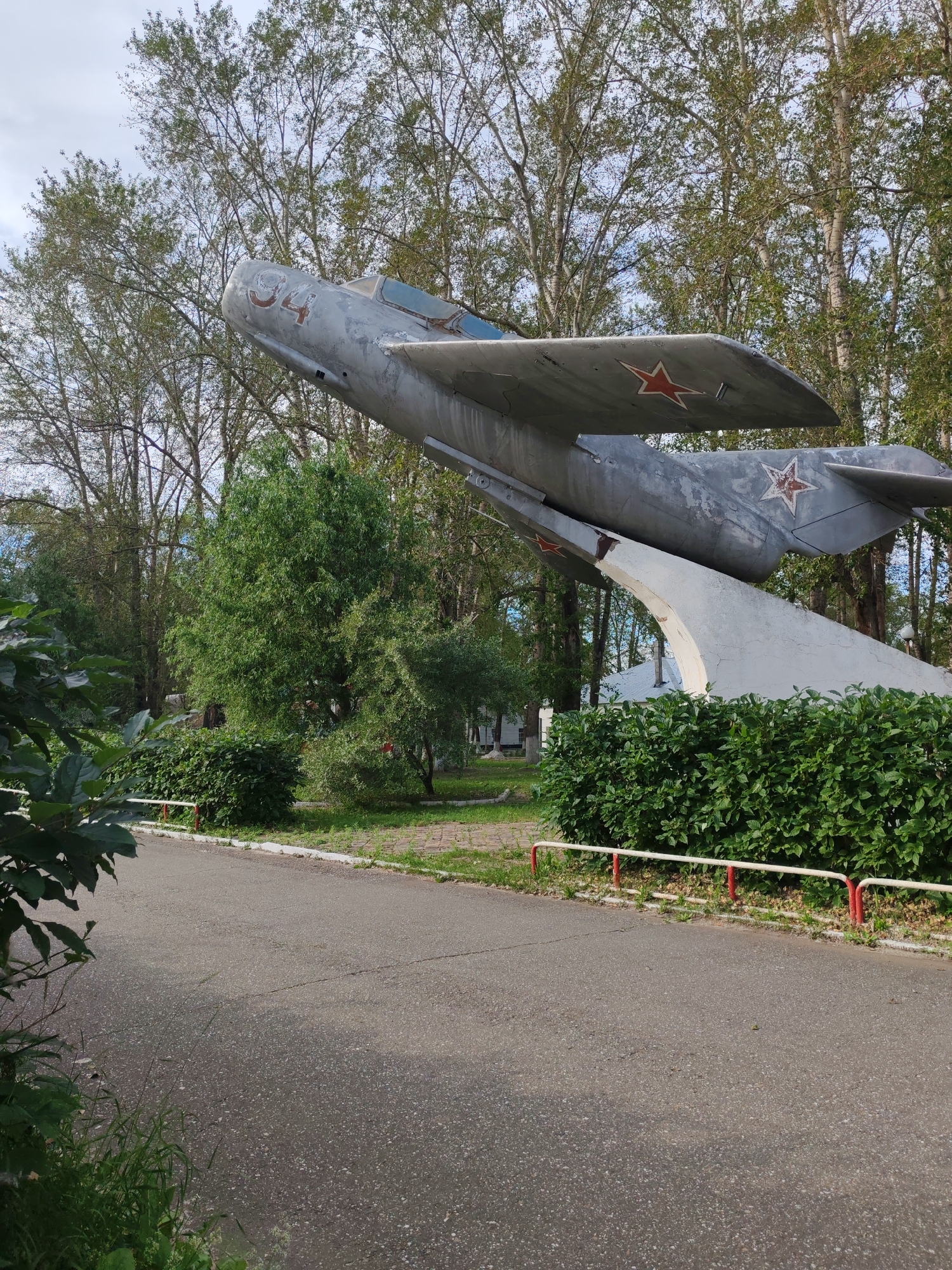 This media file shows a weathered, gray fighter jet monument displayed outdoors in a park-like setting. The jet is tilted upwards, supported by a concrete structure. Red stars are visible on the wings and tail of the aircraft. The number "04" is painted on the front of the fuselage.
The scene is surrounded by lush green trees and bushes, suggesting a natural environment. A paved path runs in front of the monument, bordered by a low fence with red and white posts. The ground is covered in grass and some patches of pavement. In the background, glimpses of buildings can be seen through the trees.
The lighting and the presence of shadows suggest it is daytime, likely overcast or with dappled sunlight filtering through the trees. There is no discernible activity taking place; the scene appears to be a static display. The overall impression is one of a memorial or historical exhibit, situated in a public outdoor space in Сокол, Russia. The monument itself shows signs of age, with chipped paint and wear, indicating it has been on display for some time.