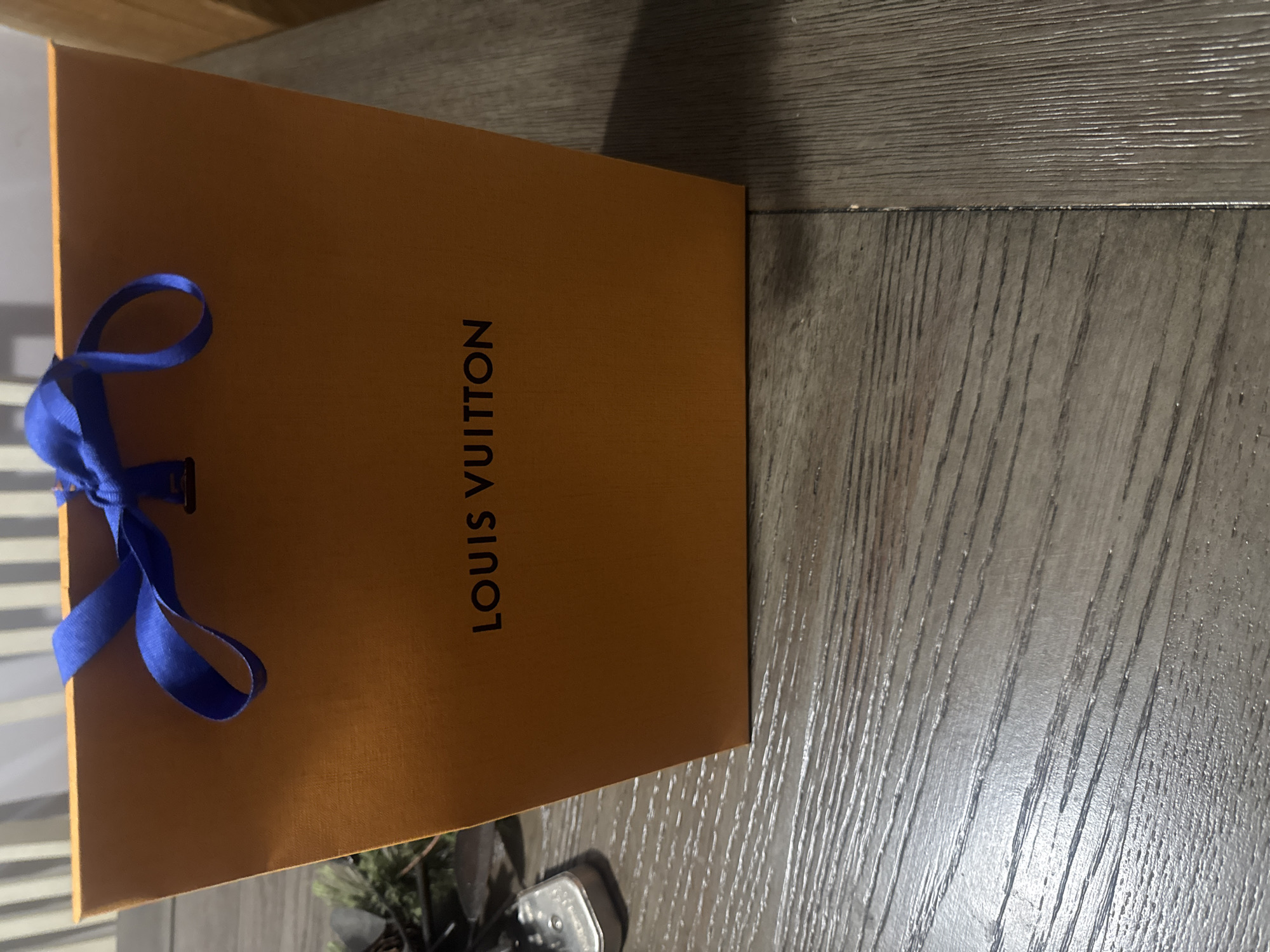 The image displays a vibrant orange Louis Vuitton shopping bag, positioned horizontally across the frame with its printed side facing the viewer. The brand name "LOUIS VUITTON" is visible in black capital letters, printed vertically along the length of the bag. A bright blue ribbon is tied into a bow, situated towards the top-left edge of the bag. The bag rests on a textured wooden surface, characterized by a distinct wood grain and horizontal plank lines, which covers the majority of the background and foreground to the right and bottom of the bag. On the far left, a blurred background element with horizontal striations suggests either a window with blinds or a wall paneling. A small, dark, indistinct object, possibly a potted plant or piece of furniture, is partially visible in the bottom-right corner. The scene is illuminated by soft, ambient indoor lighting, casting subtle shadows from the bag. The photograph is a still life, primarily showcasing the luxury brand's packaging.