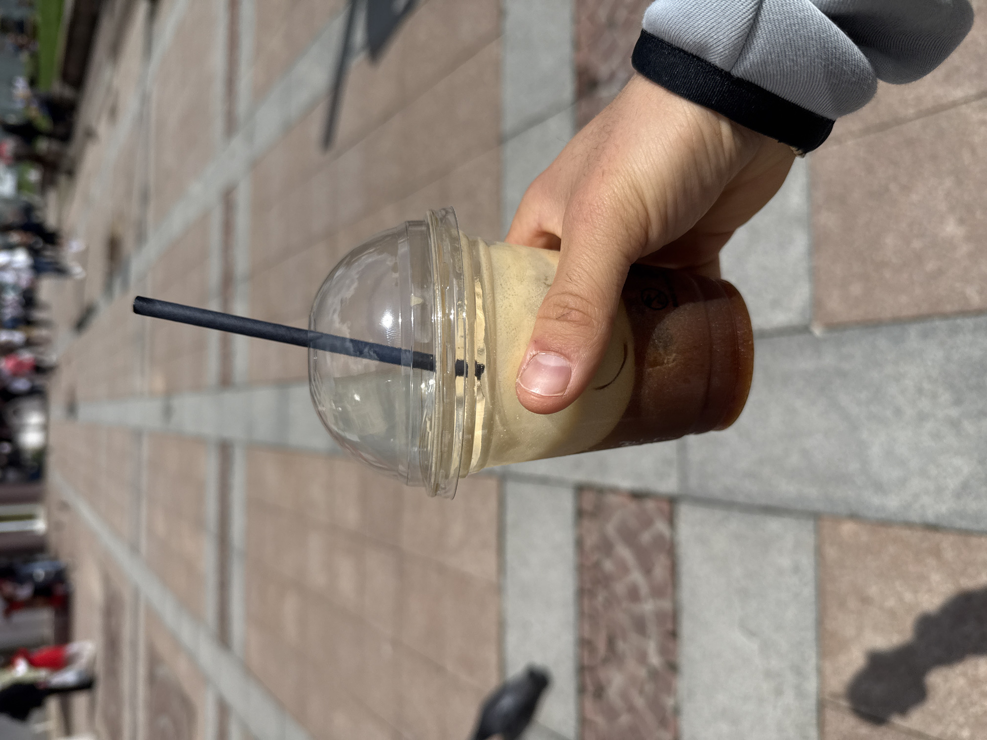 A right hand holds a clear plastic cup containing a layered iced coffee drink. A black straw is in the drink. The hand has on a gray and black jacket sleeve. The background shows a brick-paved walkway with people in the distance. The location is Gabrovo, Bulgaria.