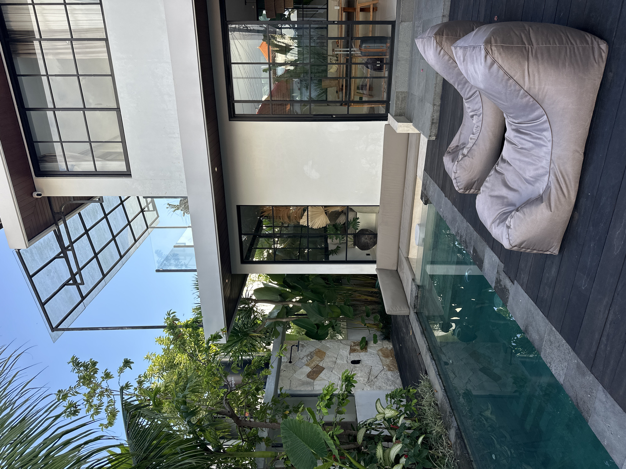 The image depicts the exterior of a modern residence in North Kuta, Indonesia. The architectural style is characterized by clean lines, a flat roof, and large glass windows with black frames. The building is primarily white, with wood accents and a glass canopy extending from the upper level.
A swimming pool runs alongside the building, partially lined with wood decking that also features two large, grey beanbag-style chairs. A built-in beige sofa lines part of the pool. Lush tropical foliage, including palm trees and other greenery, surrounds the area. A stone wall with a shower fixture is visible amongst the plants. The sky is clear and blue.
There are no people visible in the scene. The setting suggests a luxury residence with a focus on indoor-outdoor living.
