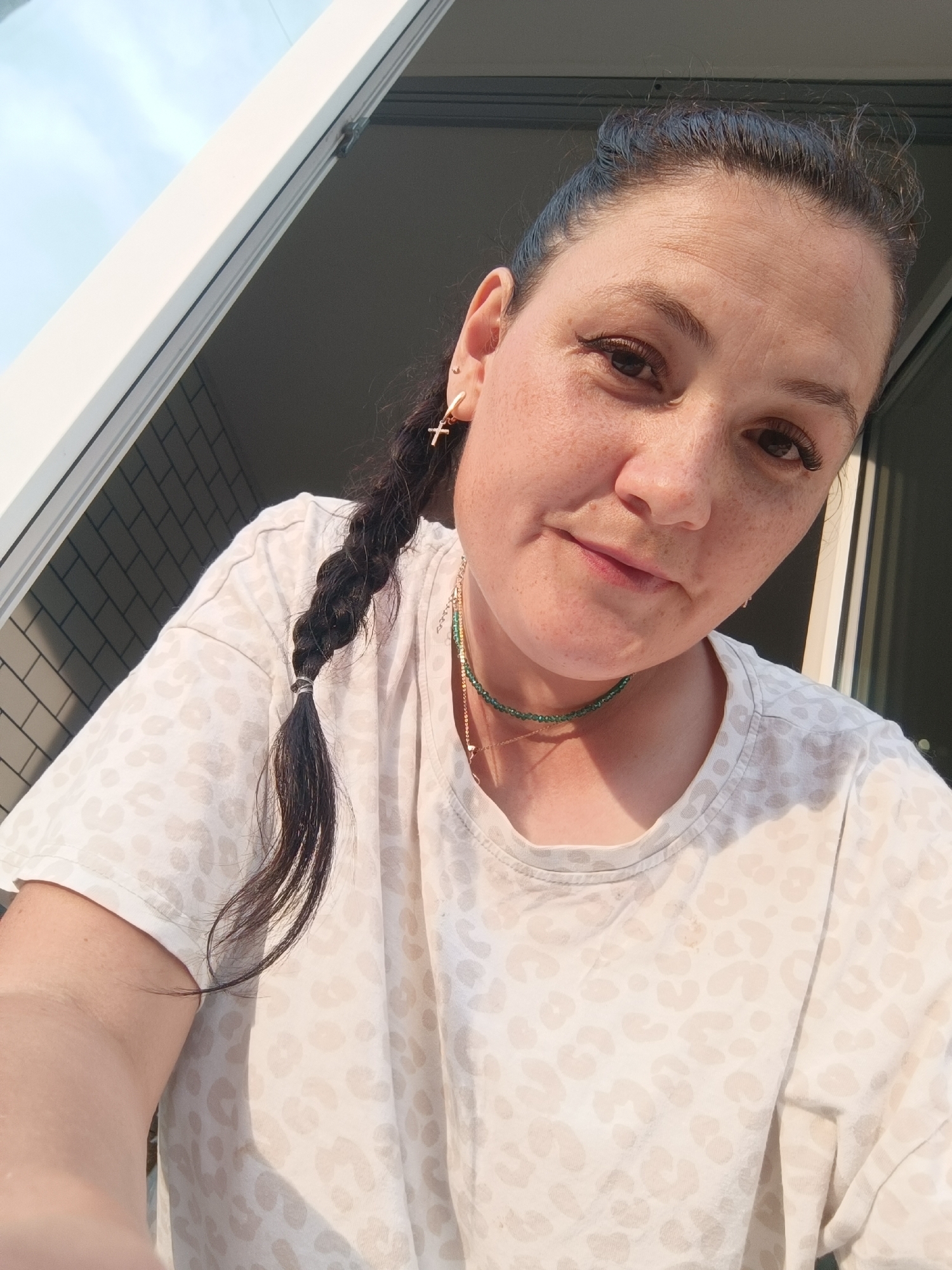 In this close-up, high-angle selfie, a Caucasian woman with dark hair styled in a braid is looking at the camera. She is wearing a light-colored t-shirt with a subtle leopard print pattern and a green beaded necklace. A small cross pendant hangs from a gold chain around her neck. She has a gold earring in her left ear, featuring a small cross charm. The background shows an outdoor scene with a white railing and what appears to be a brick wall. The lighting suggests it is daytime, with sunlight casting shadows and highlighting the woman's features, including freckles on her face. The image was taken in the city of Chaikovsky, Russia.