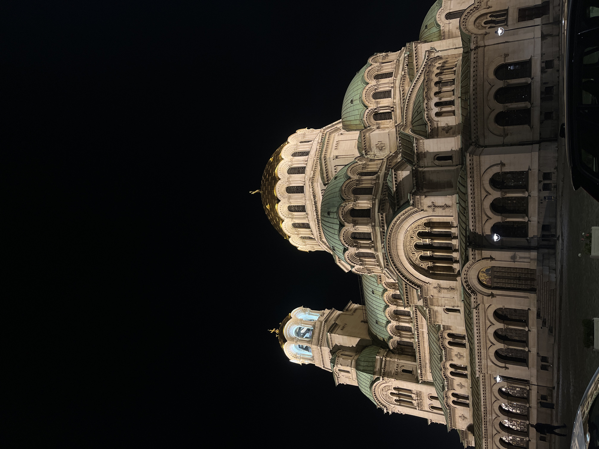 The image captures a dramatic nighttime view of the Alexander Nevsky Cathedral in Sofia, Bulgaria. The monumental Neo-Byzantine structure is brilliantly illuminated against a completely dark, clear sky, highlighting its intricate architectural details. The building's facade, likely constructed of light-colored stone or marble, features a grand central golden dome, which glows brightly, and several smaller, distinctive green domes that cap various sections of the cathedral.
The cathedral exhibits multiple tiers adorned with arched windows, decorative cornices, columns, and elaborate carvings. The artificial lighting emphasizes the textures and depth of these ornamental elements, creating a striking contrast with the black background. On the far right side of the image, a portion of a dark vehicle, possibly a car or bus, is visible. Near the base of the cathedral, a faint silhouette of a person can be observed, providing a sense of scale to the immense building.
The scene takes place at night, confirmed by the dark sky and the presence of artificial illumination on the building. There are no visible weather conditions like clouds or precipitation, indicating a clear night. The architectural style, including the domes and ornate design, is characteristic of Eastern Orthodox cathedrals, specifically identifying it as the iconic Alexander Nevsky Cathedral in Sofia. No discernible text is visible within the image.