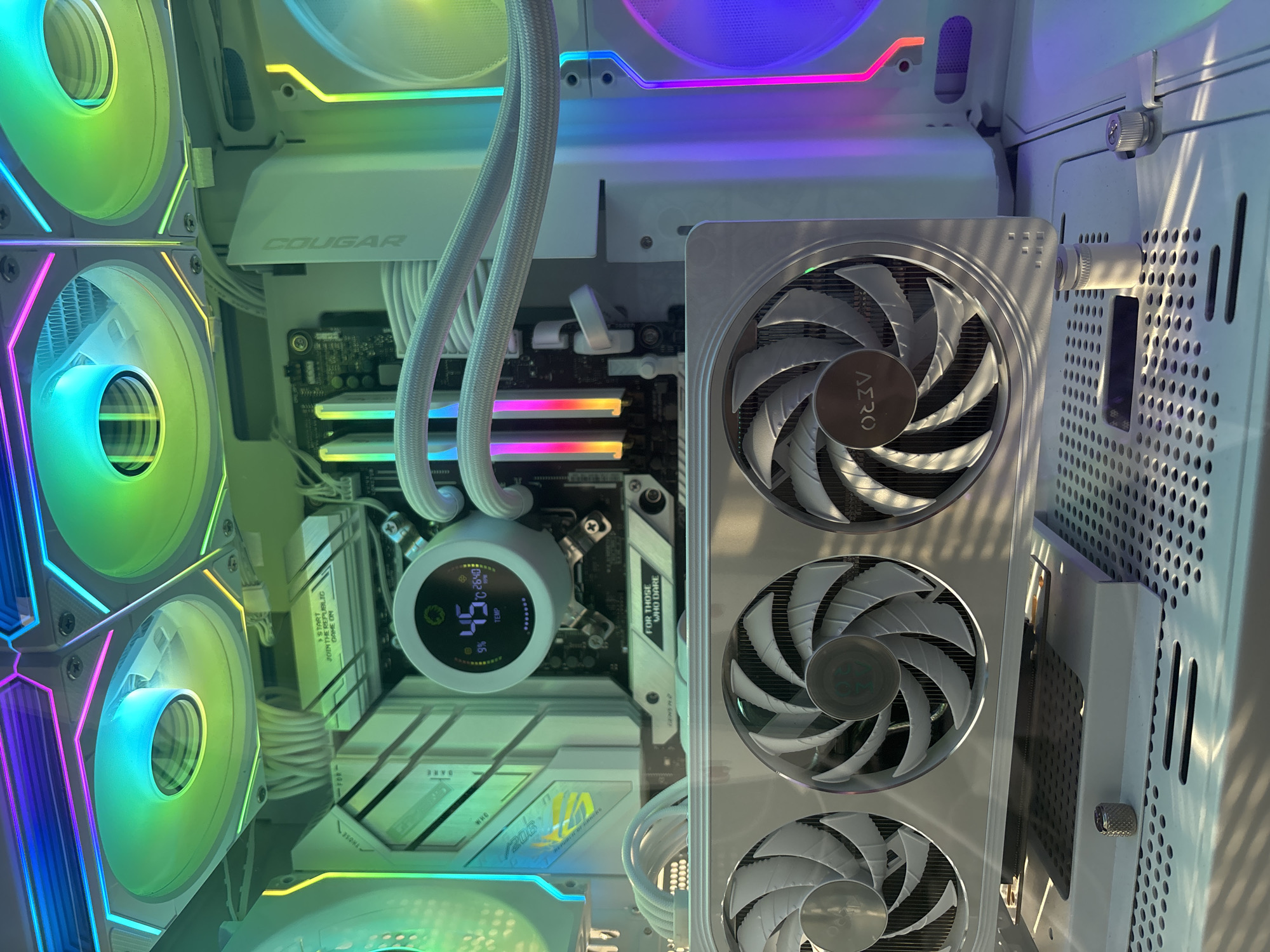 This is a close-up, high-angle shot of the interior of a white computer tower. The computer is lit by a variety of RGB lighting effects, casting colorful hues of green, blue, pink, and purple across the components.
The main subjects are the internal computer parts, including multiple fans with RGB lighting, a CPU cooler with a digital display showing "45 TEMP" and "9%", RAM modules with RGB lighting, and various circuit boards. The brand "COUGAR" is visible on a white component. Another visible brand logo is "AERO" on the central fan assembly. Text is also visible on some components, including "FOR THOSE WHO DARE" and "START JOIN THE REPUBLIC GAME ON".
The scene appears to be well-lit and organized, suggesting a functional and possibly custom-built PC. There are no people or external environmental cues to indicate the time of day, weather, or specific location. The focus is solely on the intricate details of the computer hardware and its lighting.