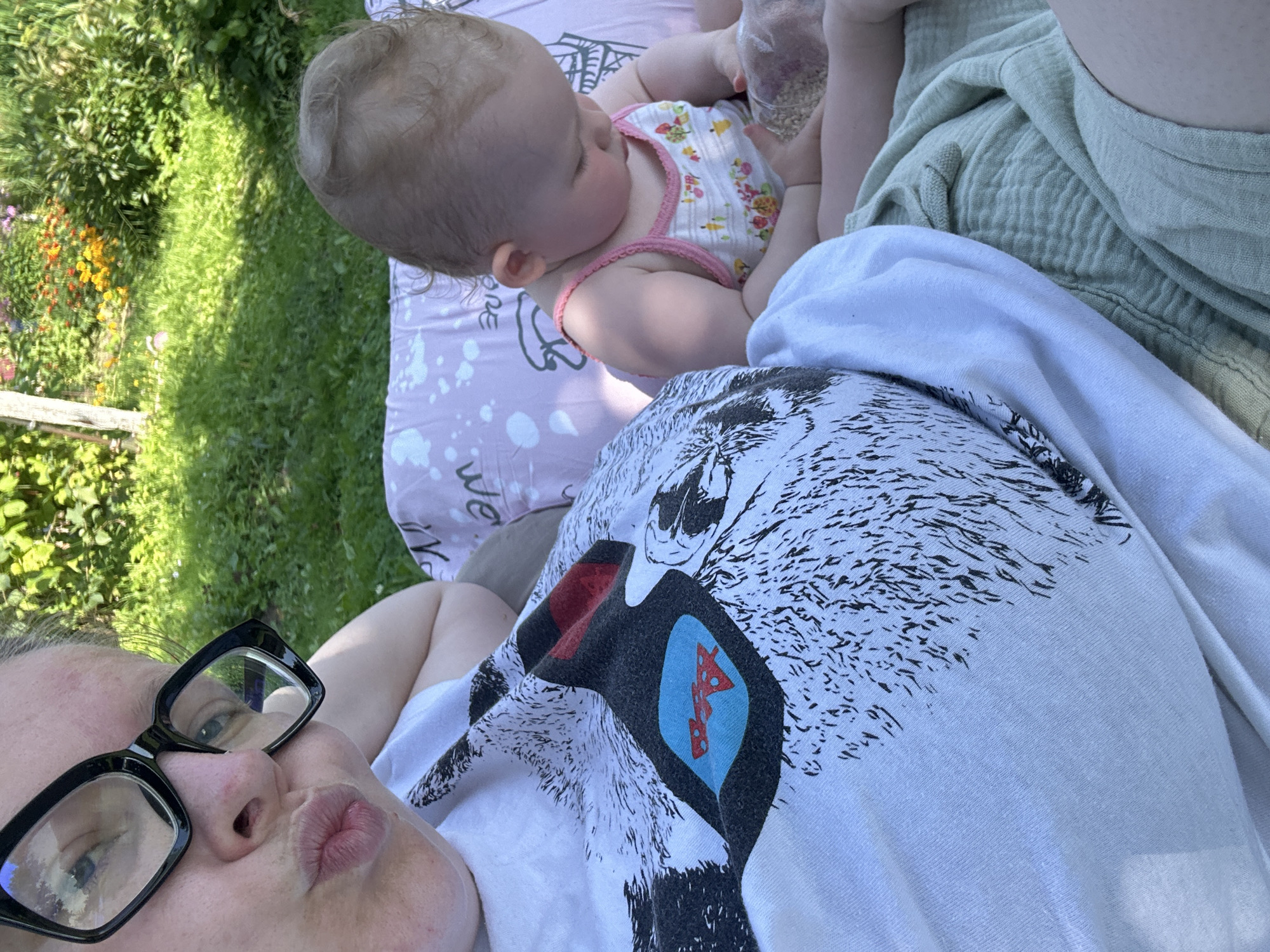 A woman with glasses is taking a selfie outdoors in what appears to be a yard with green grass and flowers in the background. She is wearing a white t-shirt with a graphic print and is puckering her lips. Next to her, a baby in a floral tank top is sitting and playing with a clear plastic bag filled with small objects. The location is not specified in Vereiki, Russia.