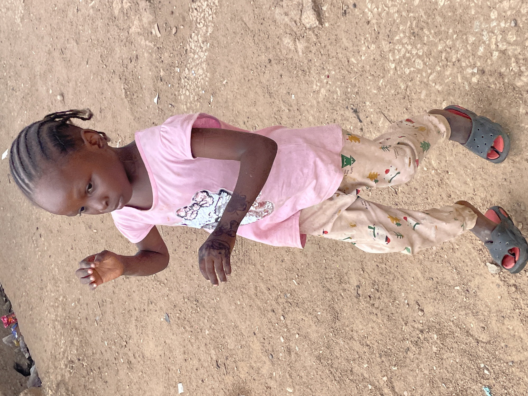 A young Black child lies prone on a tan, dusty ground in Jos, Nigeria. The child, wearing a pink shirt with a floral print and beige Christmas-themed pajama pants with black and red sandals, is looking down. The left arm has decorative dark designs on the forearm and hand. The ground is uneven with small rocks, debris, and trash visible along the left edge of the frame.