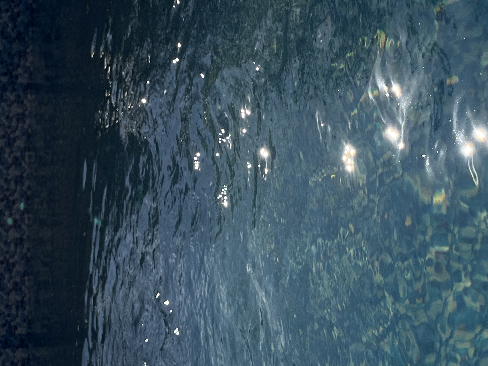 The image shows a close-up of dark, rippling water with bright highlights. The surface of the water is textured, with small waves and disturbances creating dynamic patterns. Scattered across the water are numerous small, bright specks, which appear to be reflections of light. These highlights are concentrated in several areas, forming clusters of shimmering points. The water itself is a deep, dark blue, with hints of darker tones suggesting depth or shadows. In the lower right portion of the image, the water's surface seems to distort the view of what lies beneath, revealing patches of muted colors and shapes that could be rocks, sand, or other submerged objects. The left side of the image features a darker, more opaque area that appears to be the edge of something, possibly a wall or a submerged structure, with a granular texture. The lighting suggests either sunlight or artificial light reflecting off the water's surface. No people, objects, or text are clearly identifiable within the image. The overall impression is one of abstract beauty and the play of light on water.